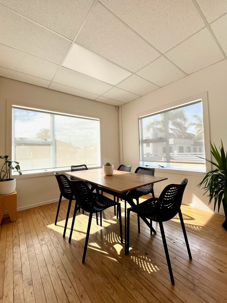 A well-lit meeting room with a rectangular wooden table surrounded by six black chairs. Large windows allow natural light to fill the space, and two potted plants add a touch of greenery. The floor is wooden, and the ceiling has white tiles.