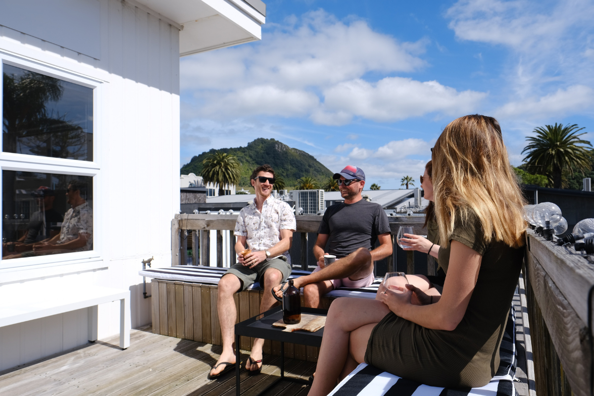 Four people sitting and talking on a rooftop terrace with a mountainous view, under a blue sky with clouds.