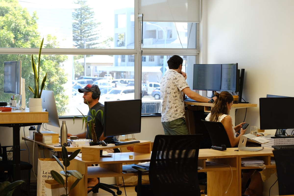 People working in a modern office with computers and plants, one man standing at a desk with dual monitors, another man sitting with headphones, and a woman looking at a smartphone.