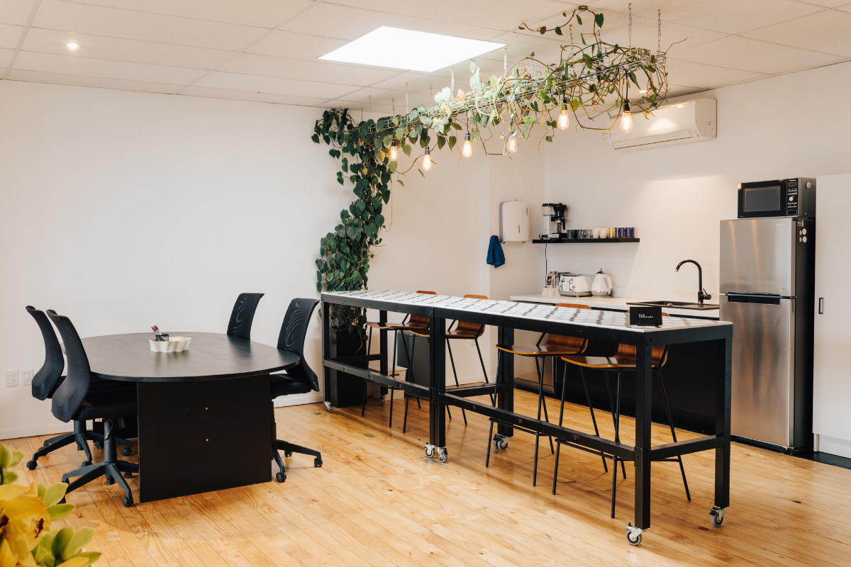 Modern office kitchen and dining area with black furniture, a long table on wheels, plants hanging from the ceiling, and stainless steel fridge and microwave. The space includes a coffee maker, shelves with cups, and a wooden floor.