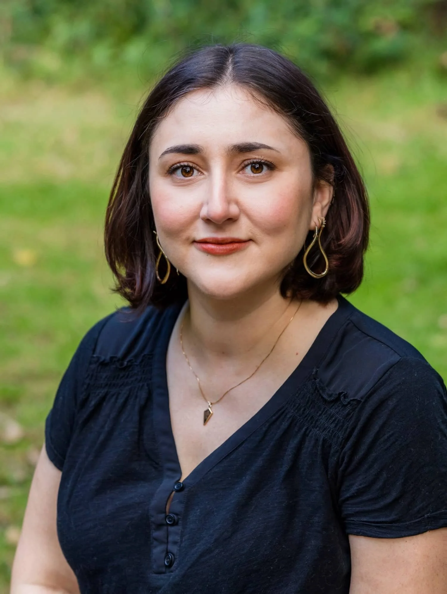 A woman with medium-length dark hair, wearing a black top, gold earrings, and a gold necklace, is outdoors with a blurred green background.