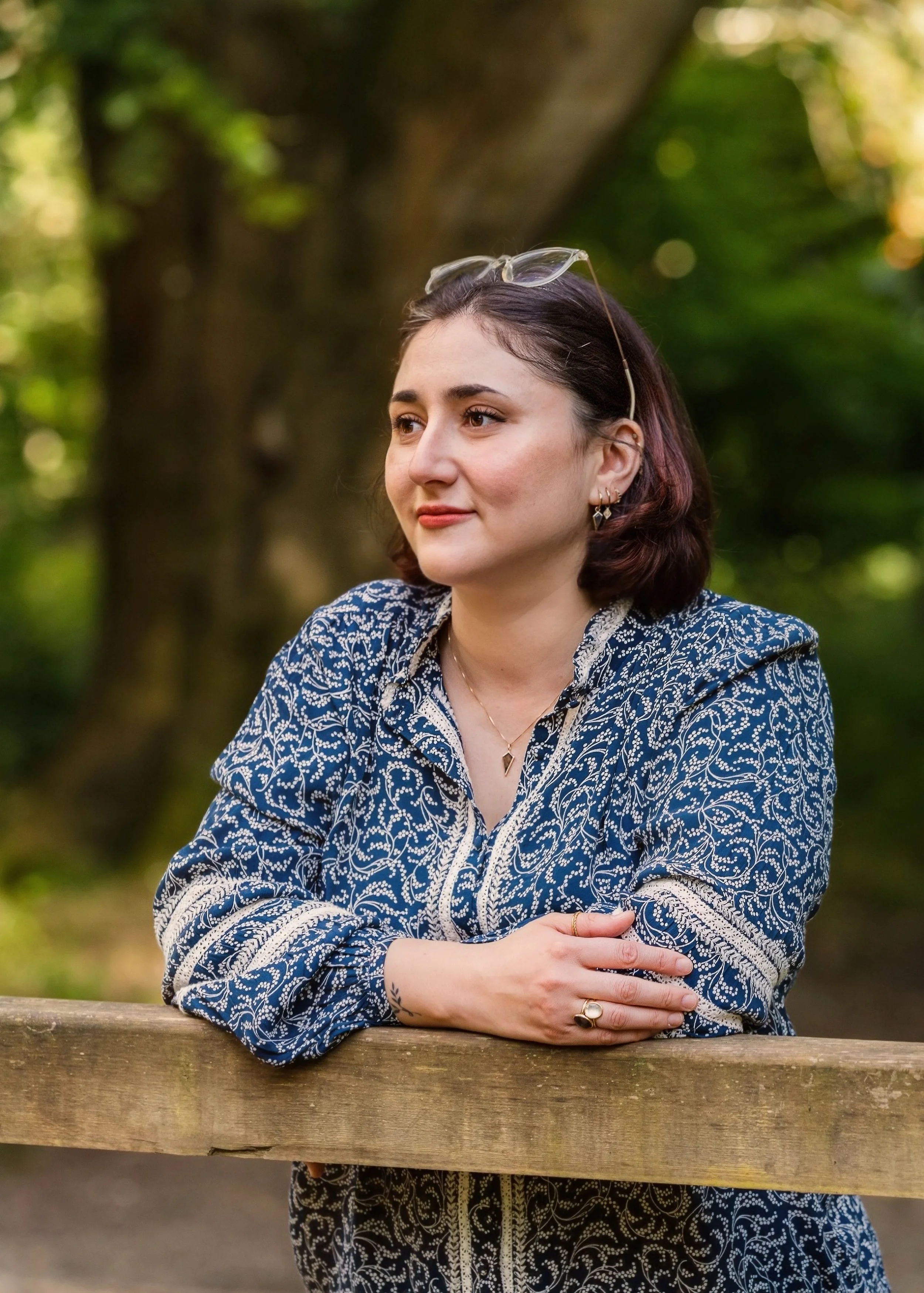 Kristina, a white woman with short dark hair, wearing glasses on her head, a patterned blue and white shirt, and gold jewelry, looking into the distance and leaning on a wooden railing outdoors with green trees in the background.