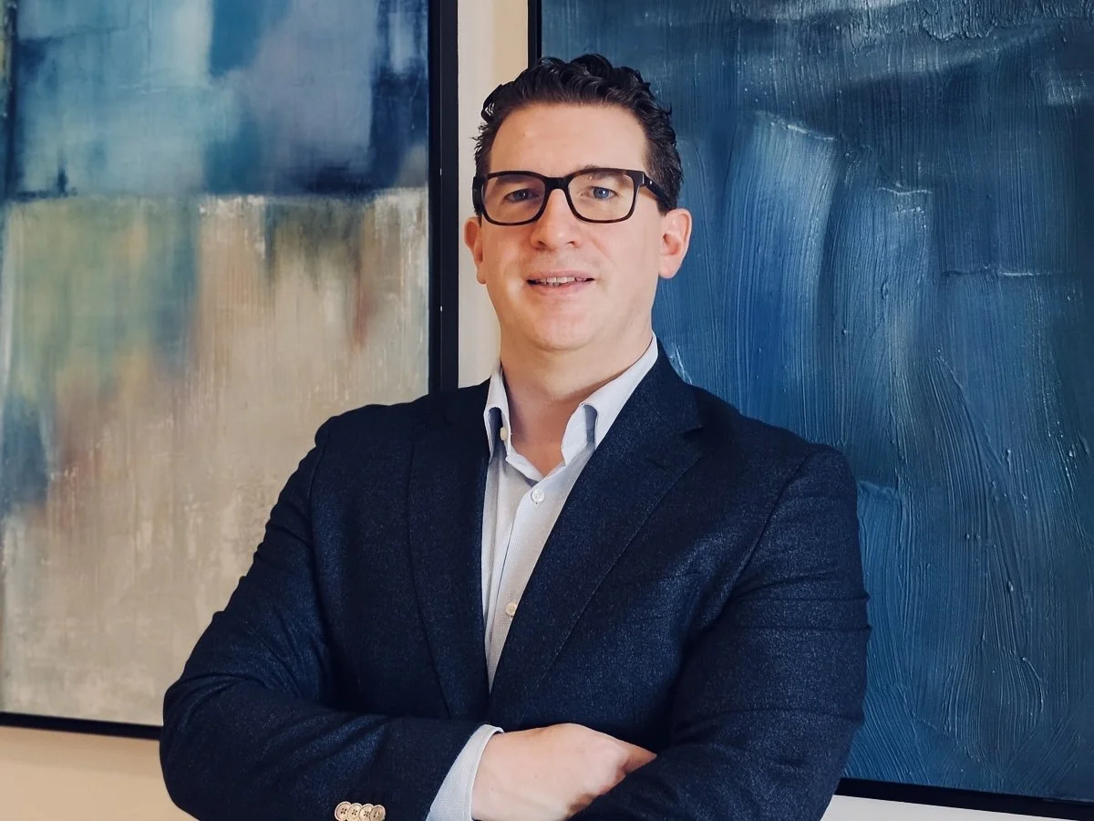 A man in a dark blazer and glasses standing in front of modern abstract artwork.