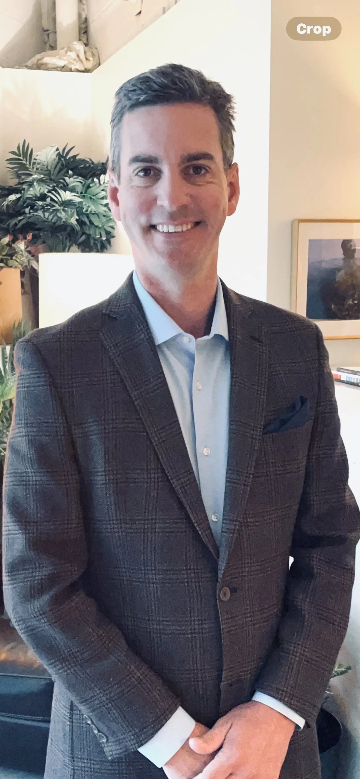 A man smiling, wearing a dark plaid blazer and light blue collared shirt, standing indoors with plants and framed art in the background.