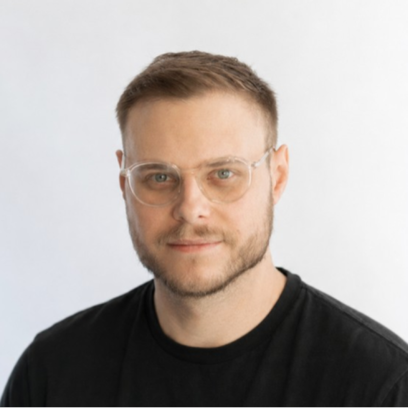 Headshot of a young man with short light brown hair, clear glasses, light beard, and mustache, wearing a black t-shirt, smiling slightly, against a white background.