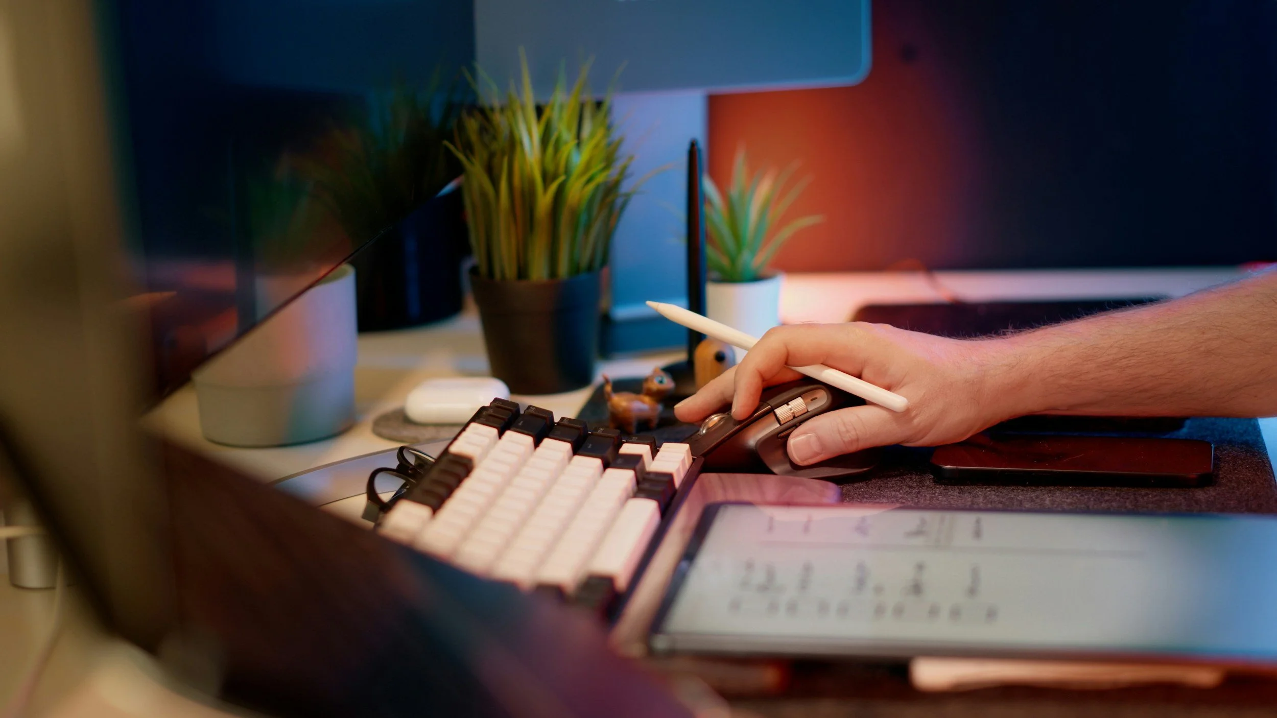 Close-up of a person's hand using a computer mouse and keyboard at a desk with potted plants and a monitor in the background.