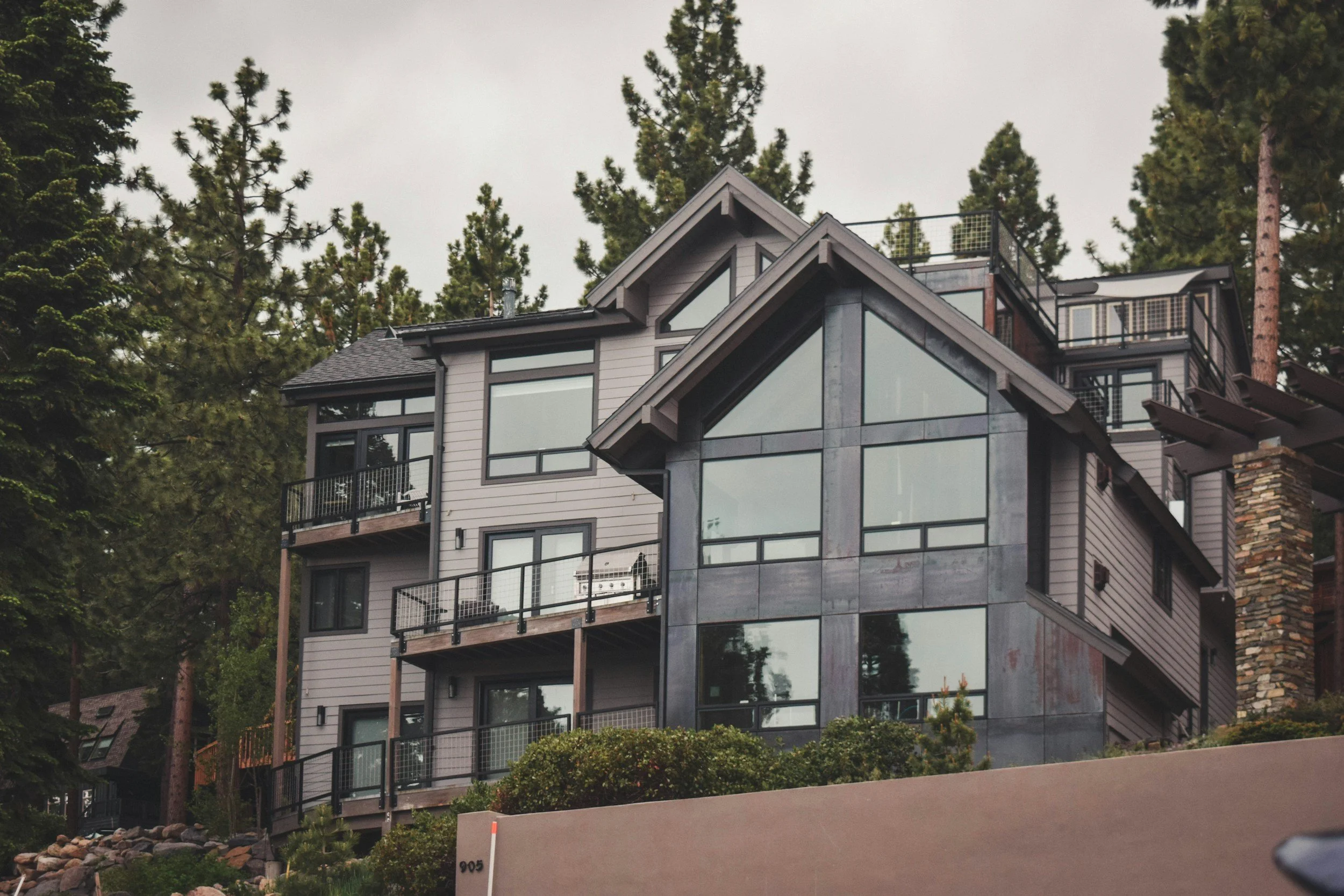 A modern multi-story house with large glass windows, gray exterior siding, and balconies, surrounded by tall pine trees in a wooded area.