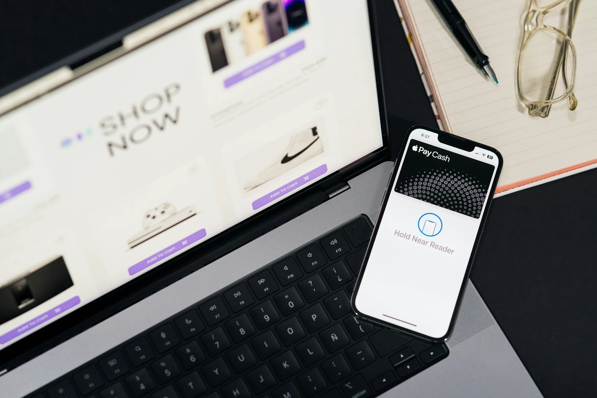 A laptop with a partially visible webpage that says 'SHOP NOW,' a smartphone displaying the Apple Pay Cash app, a black pen, a pair of glasses, and a spiral notebook on a desk.
