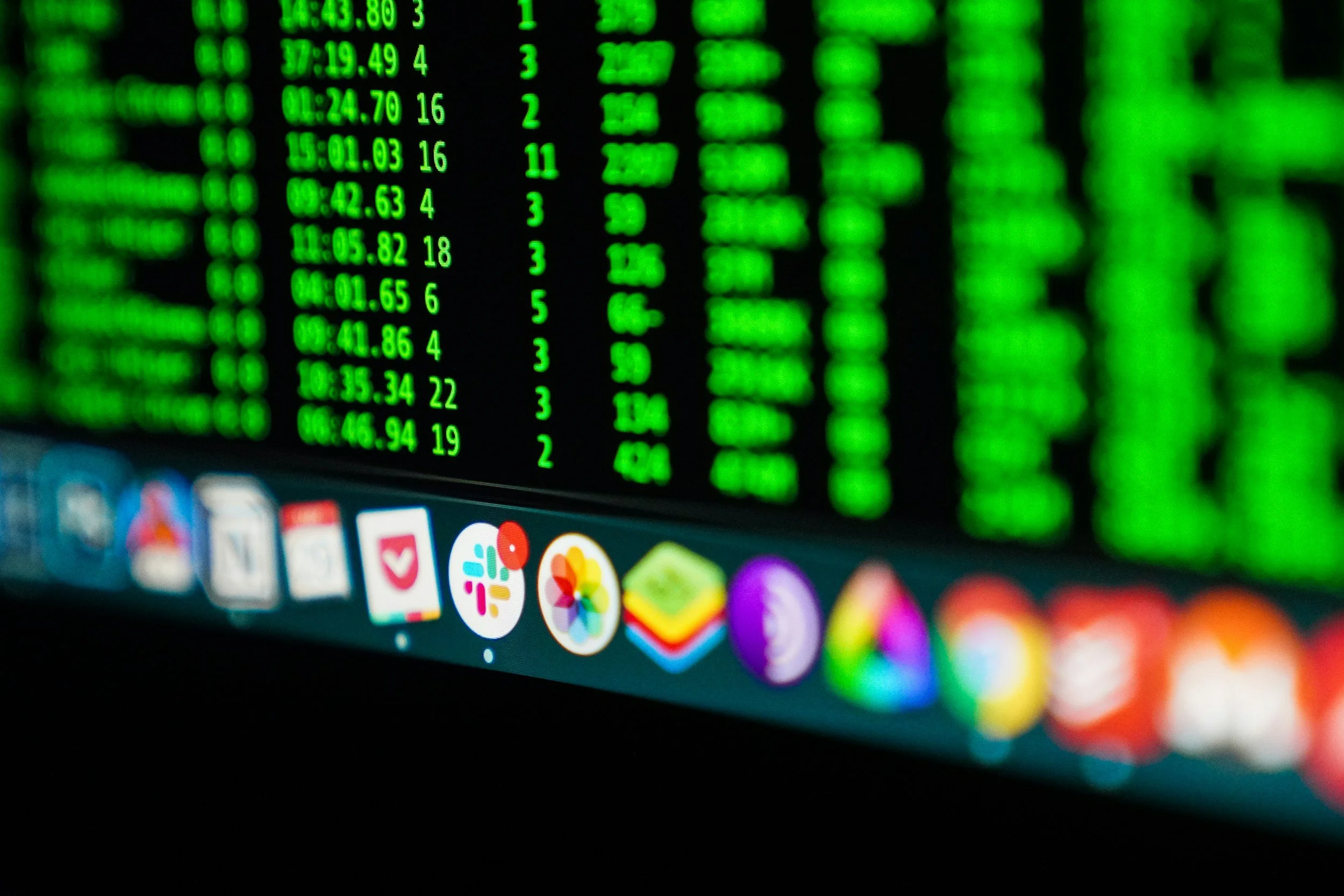 Close-up of a computer monitor screen displaying data in green text with a colorful row of app icons at the bottom.