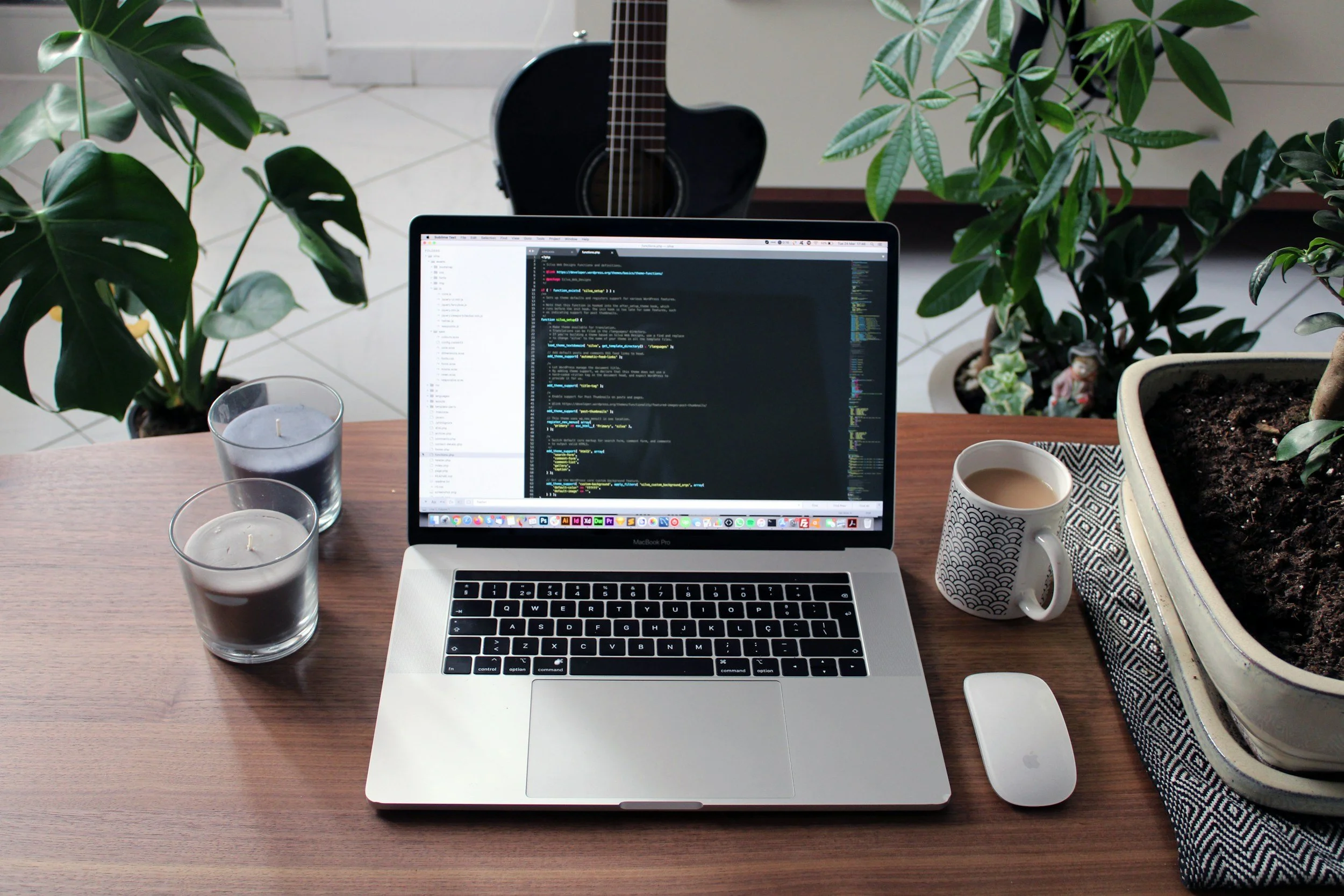 A workspace with a laptop displaying code, surrounded by three candles, a coffee mug, a mouse, and potted plants, with a guitar in the background.