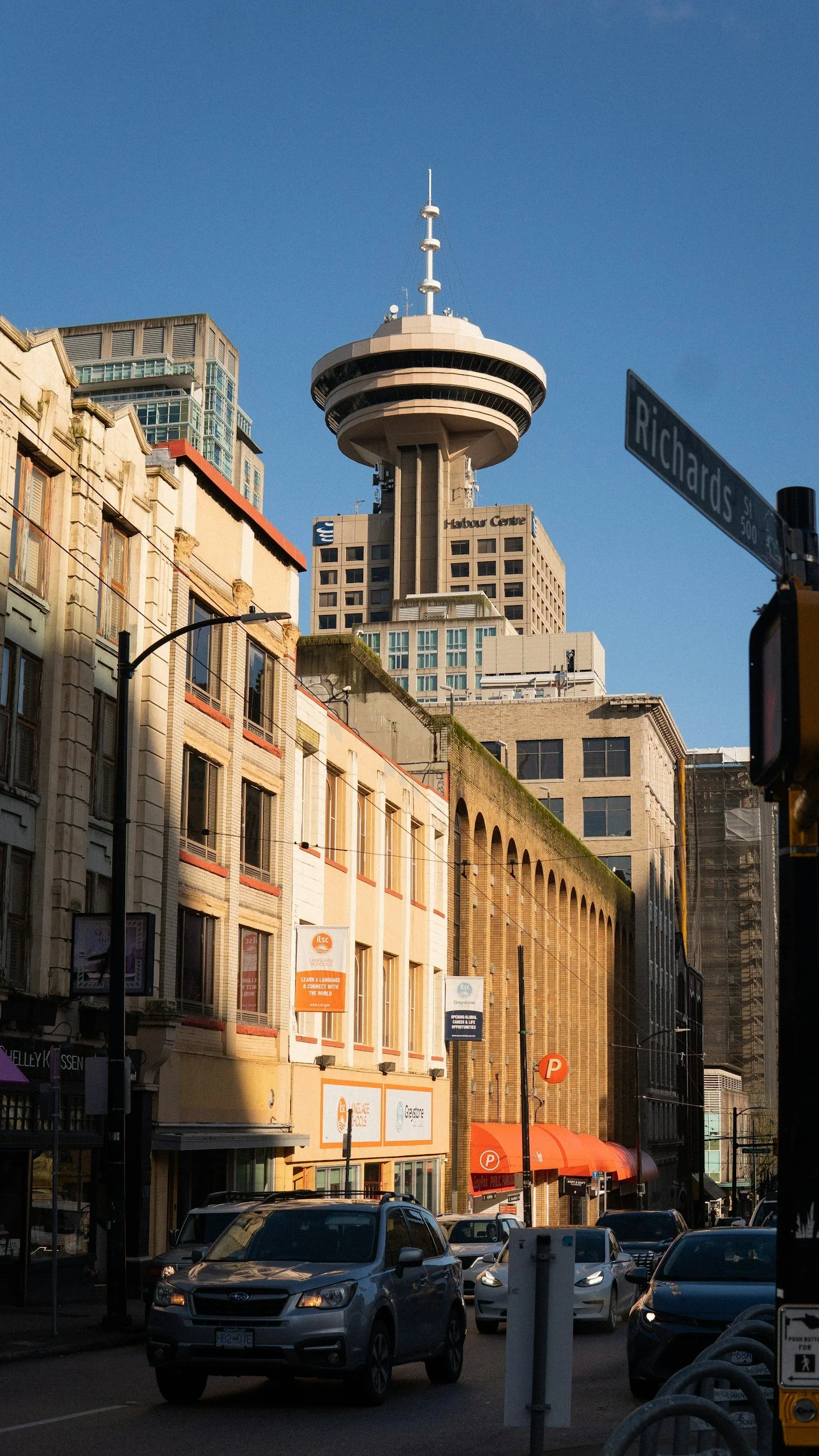Night view of a city street with cars parked and moving, multi-story buildings, and the iconic Toronto CN Tower in the background against a clear blue sky.