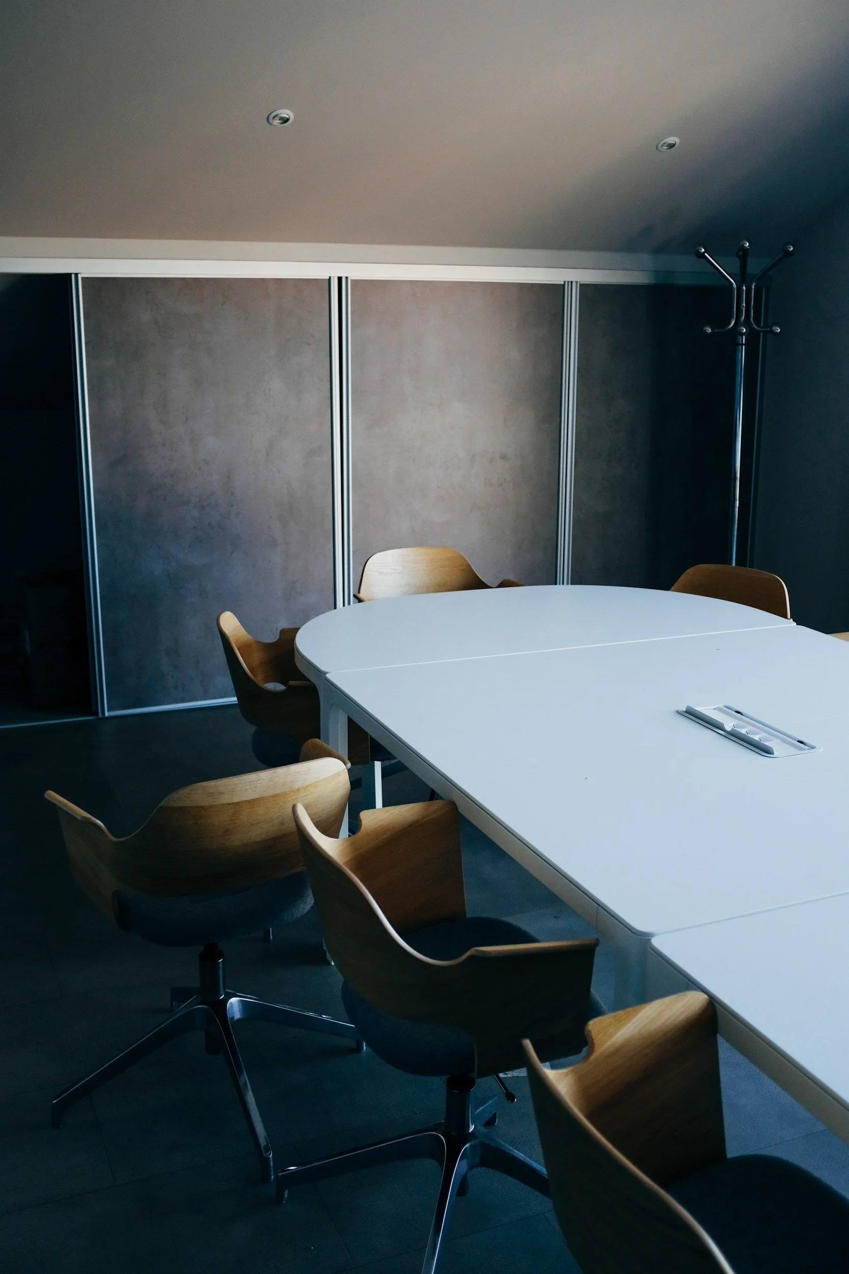 Empty office room with a white table, wooden chairs, and a grey wall with sliding panels. There is a coat rack on the right side of the room. Recessed lighting on the ceiling.