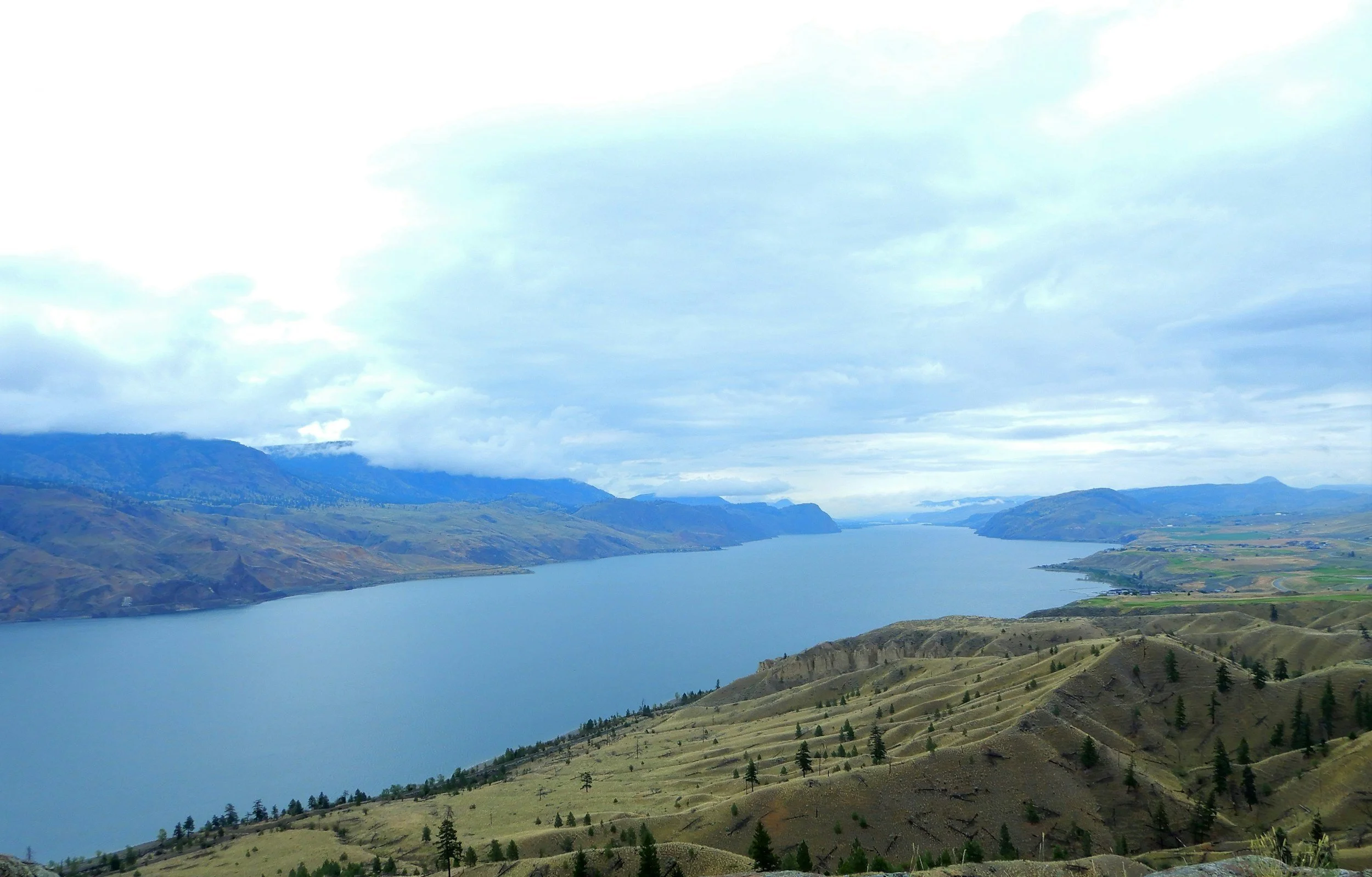 A wide river flows through a valley with rolling hills and mountains in the background. The sky is cloudy and overcast.