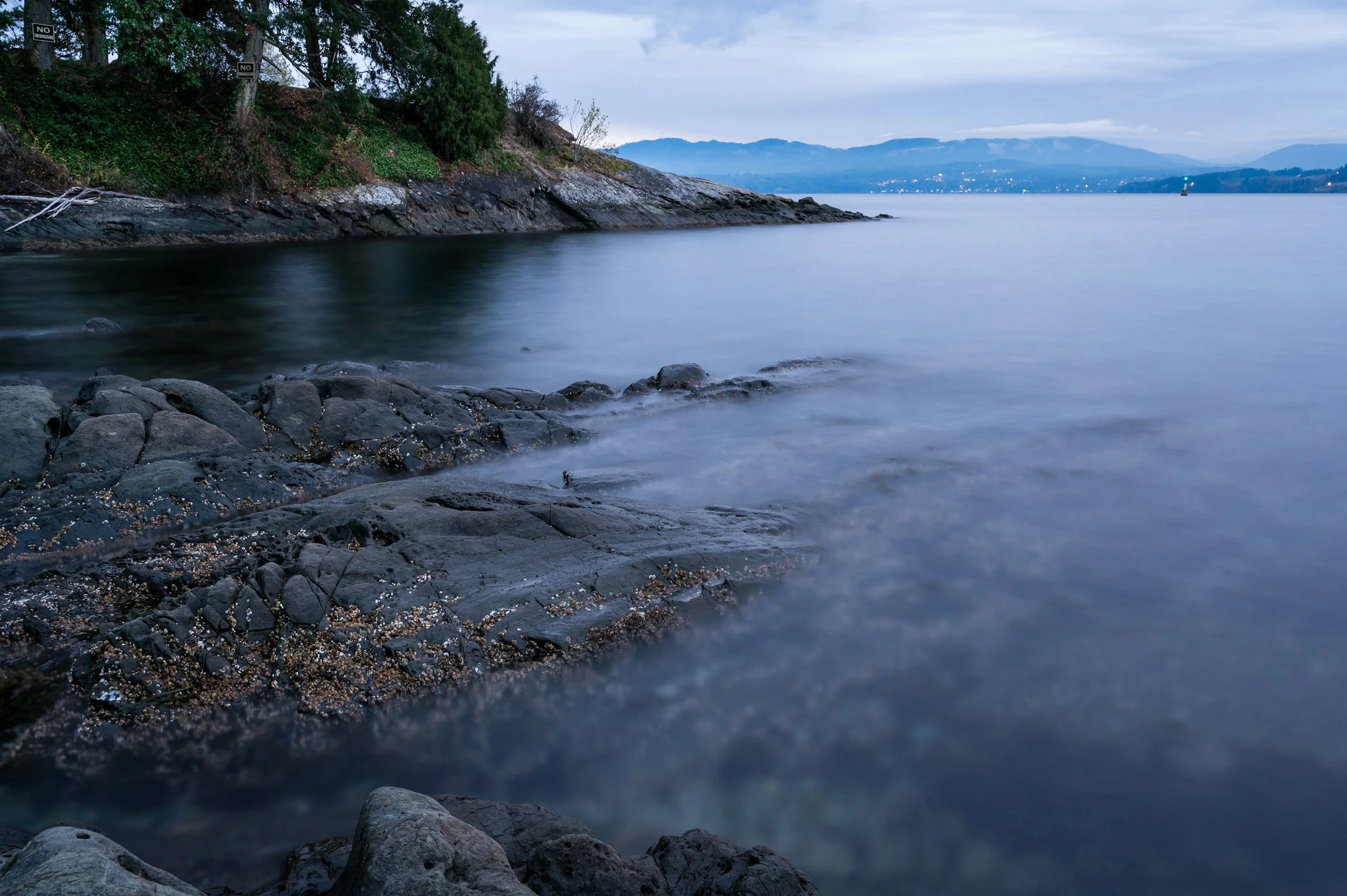A peaceful lakeside scene during twilight, with dark rocks and gentle water in the foreground, a tree-lined rocky shoreline on the left, and mountains with a cityscape in the distance under a cloudy sky.