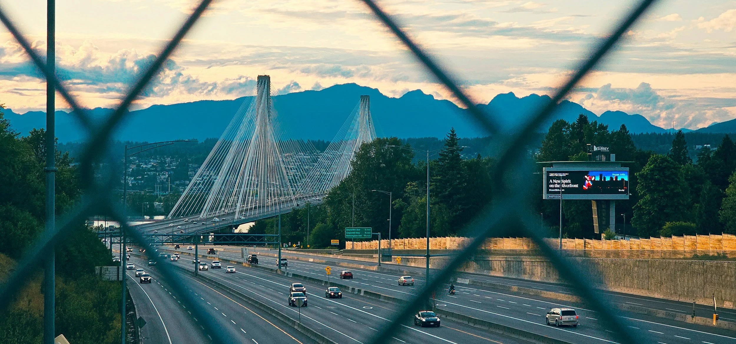 View of a suspension bridge over a highway with multiple cars and trucks, framed by a chain-link fence, with mountains and cloudy sky in the background, and a digital billboard to the right.