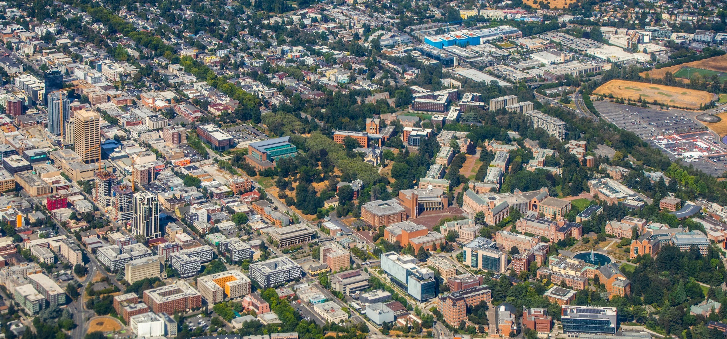 An aerial view of a city with a mix of tall office buildings, residential houses, green parks, sports fields, and parking lots.