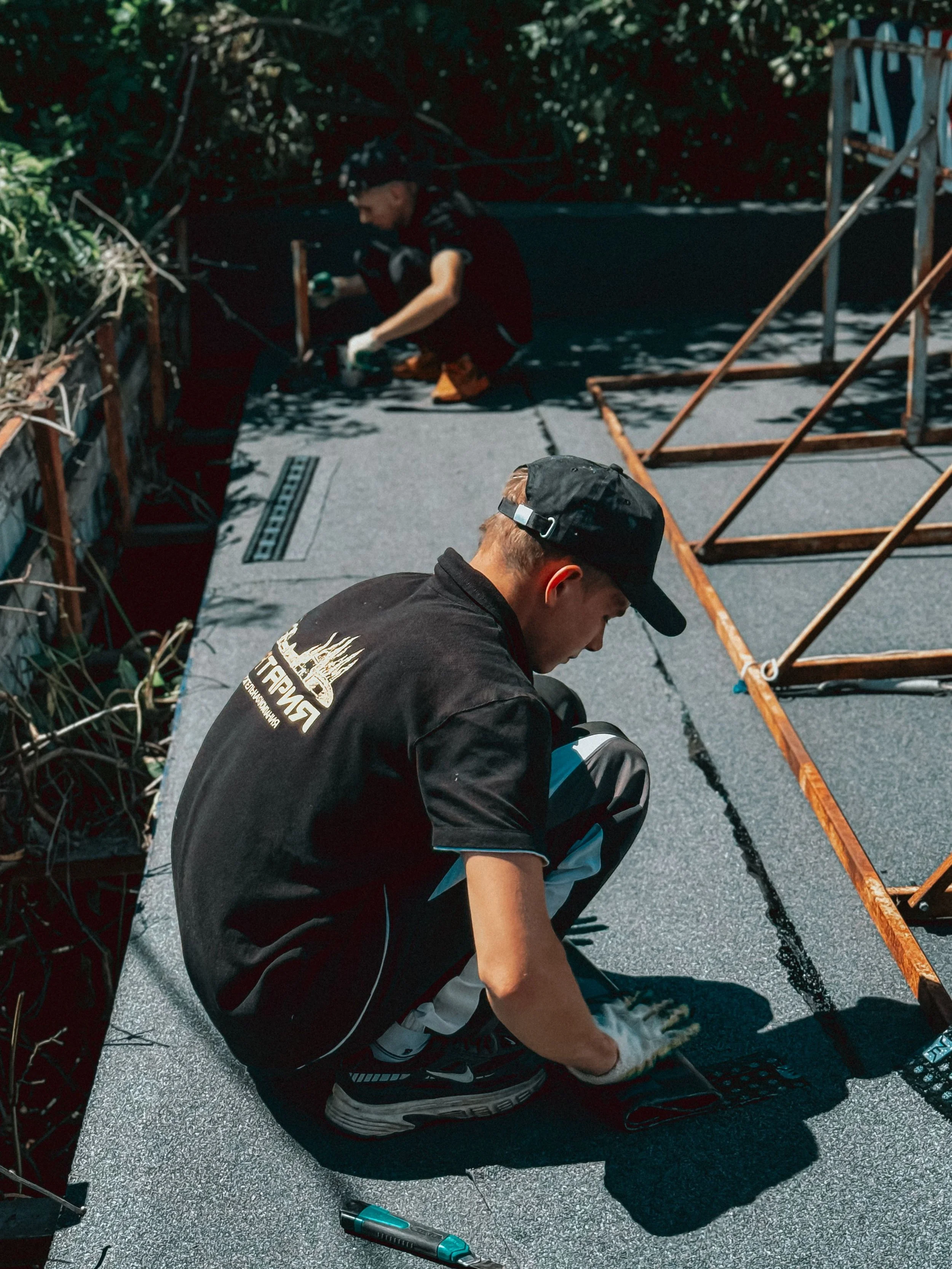 Two people working on a construction or repair project on a flat outdoor surface. One person, sitting and wearing a black cap and black shirt, is focused on a task. Another person, kneeling and wearing gloves, is also engaged in the work. There are tools and equipment around them, including an orange metal frame and a small electric device on the ground.