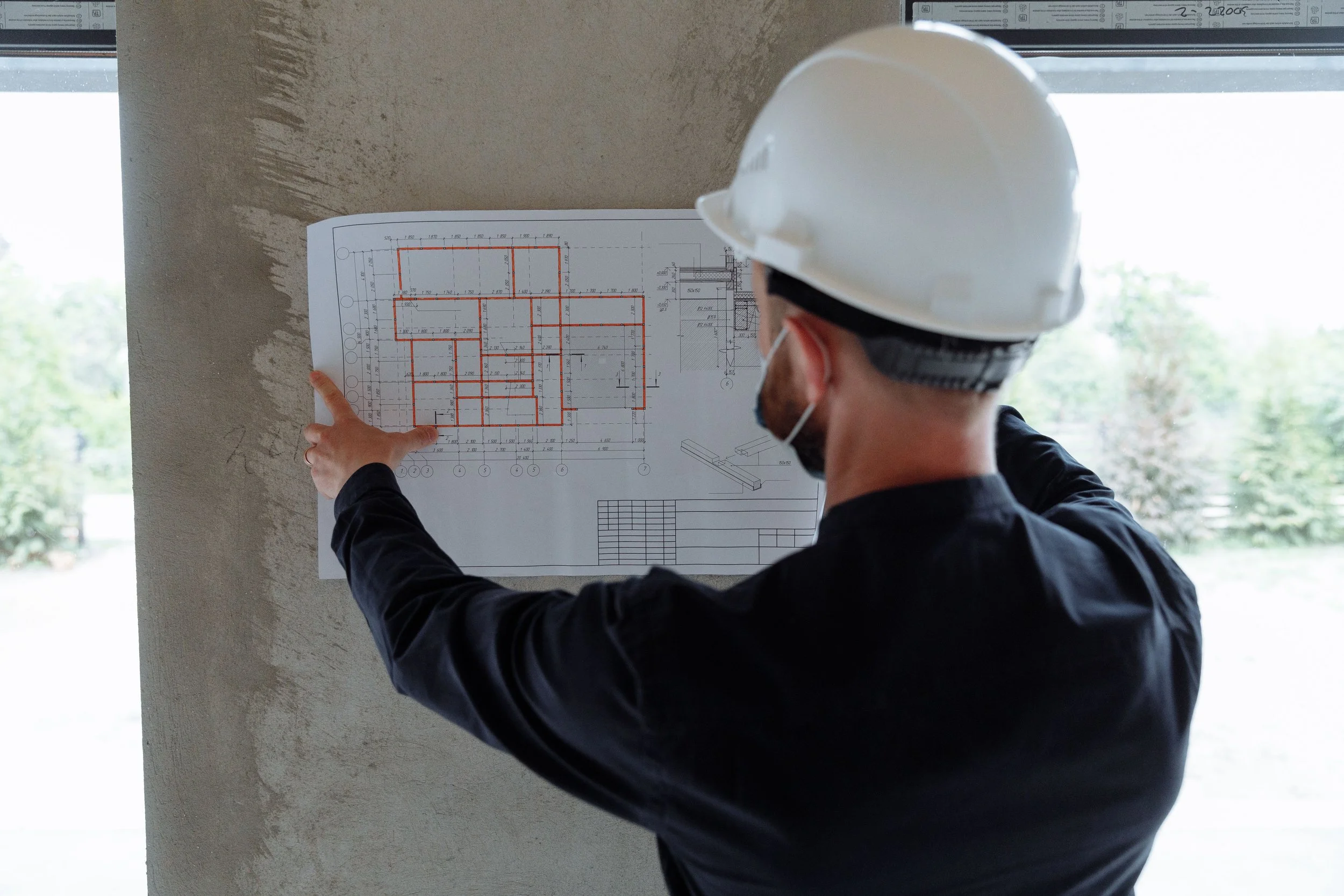 Person wearing a white construction helmet and black jacket pointing at an architectural blueprint taped on a wall.