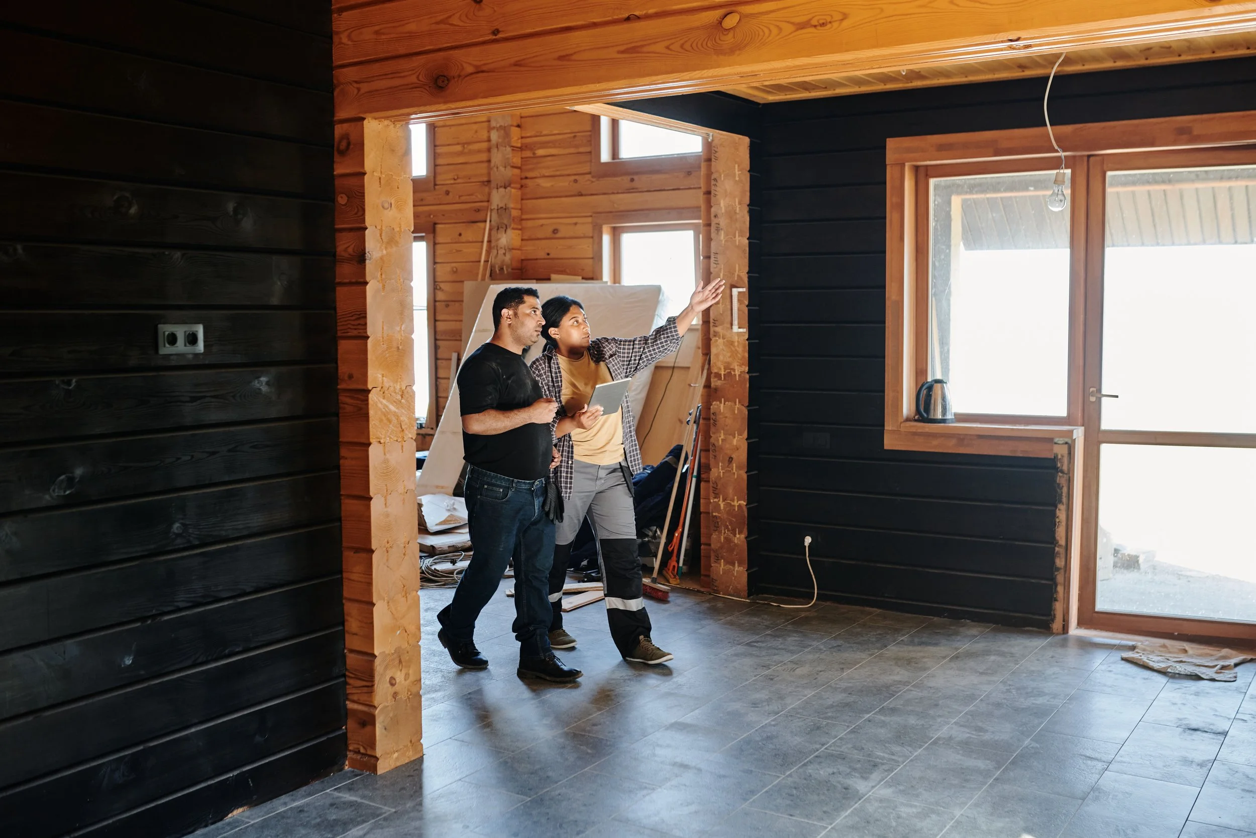 Two people, a man and a woman, inspecting the interior of a wooden house under construction, with unfinished walls, open windows, and construction materials around.
