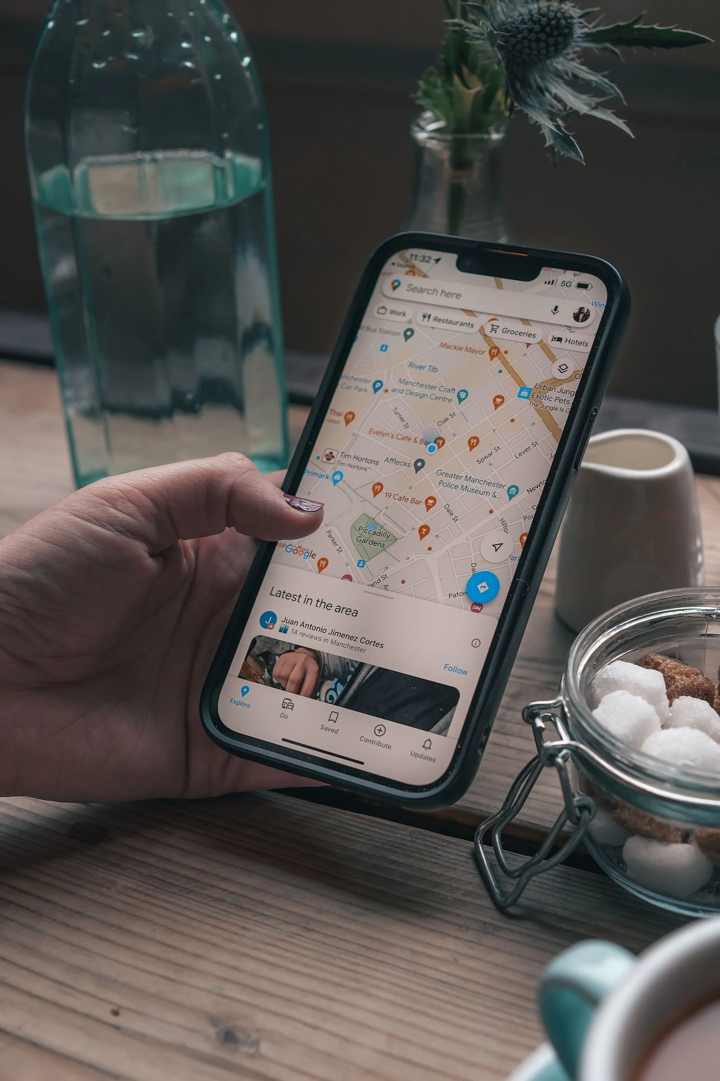 A person holding a smartphone displaying a map of Manchester, England, with various points of interest and businesses marked. On the table, there is a glass jar of sugar cubes, a small potted plant, a glass bottle of water, and a white ceramic cup.