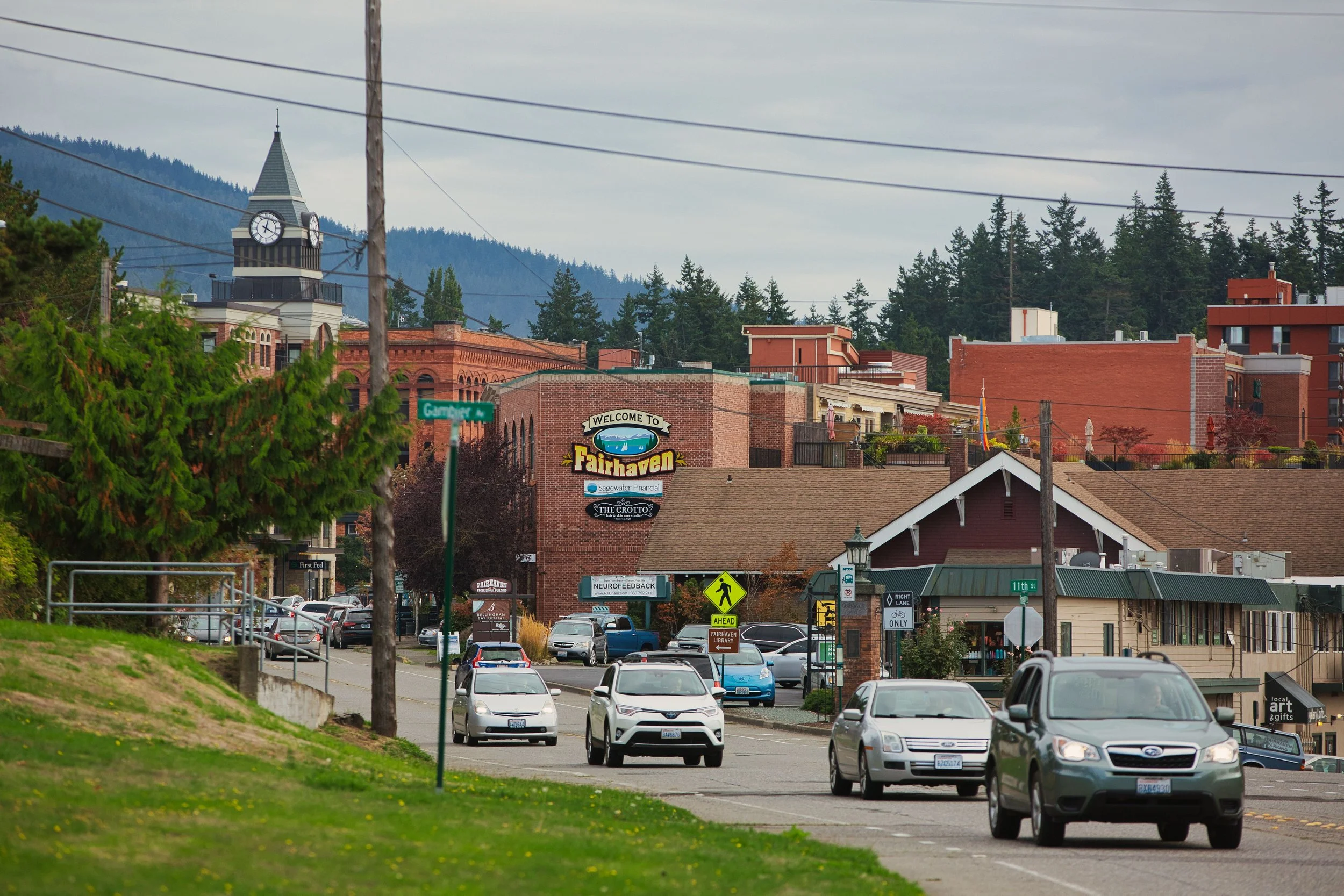 A street scene in Fairhaven with a parking lot filled with cars, a brick building with a sign that reads 'Welcome to Fairhaven,' a clock tower, several other buildings, trees, and a backdrop of mountains under a cloudy sky.