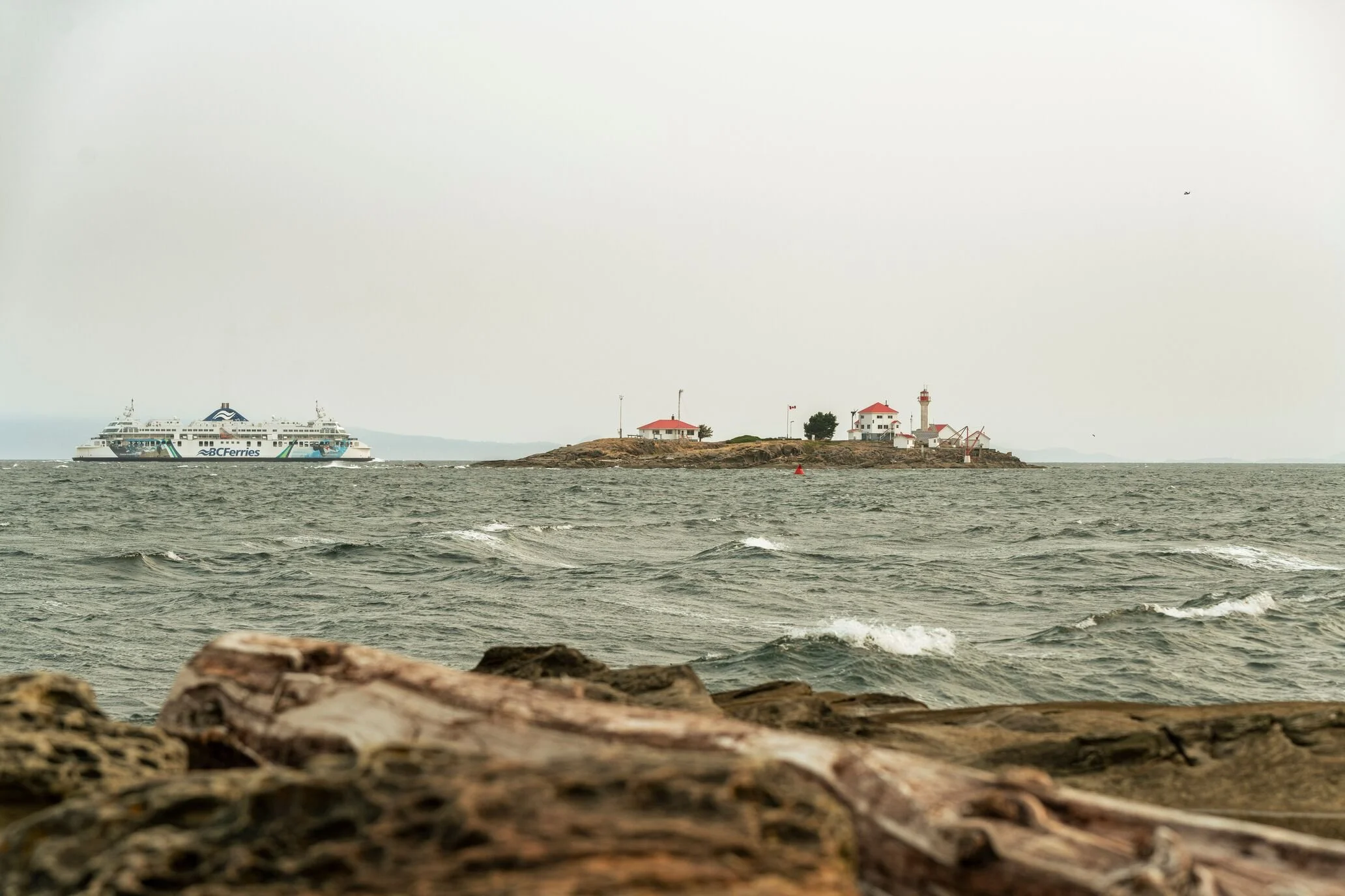 A ferry boat passing near a small island with a lighthouse and buildings, viewed from a rocky shoreline during overcast weather.