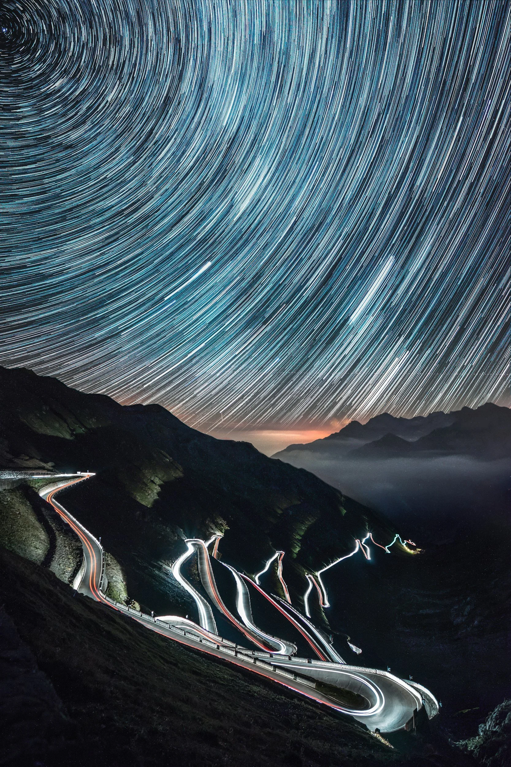Long exposure photograph of a mountain road at night with star trails in the sky above.