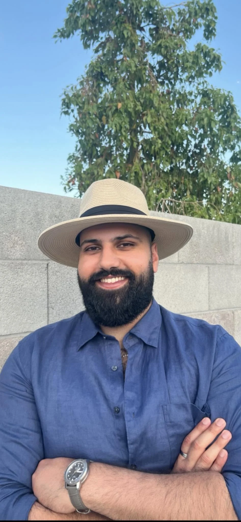 A smiling man with a beard and mustache, wearing a blue button-up shirt, a wide-brimmed straw hat, and a wristwatch, standing outdoors in front of a concrete wall with a green tree and blue sky in the background.
