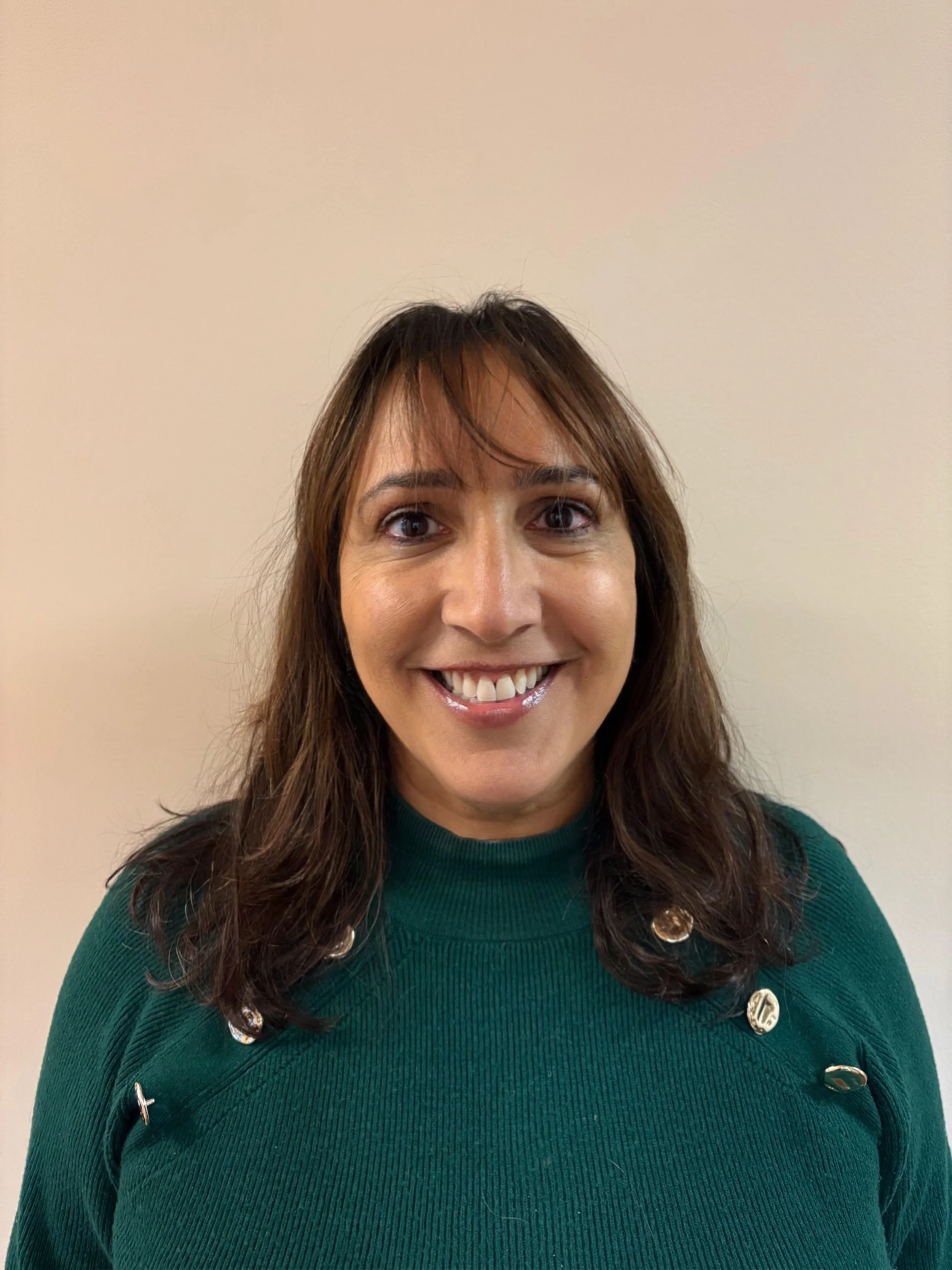 A woman with long brown hair wearing a dark green sweater with gold buttons smiling in front of a plain beige wall.