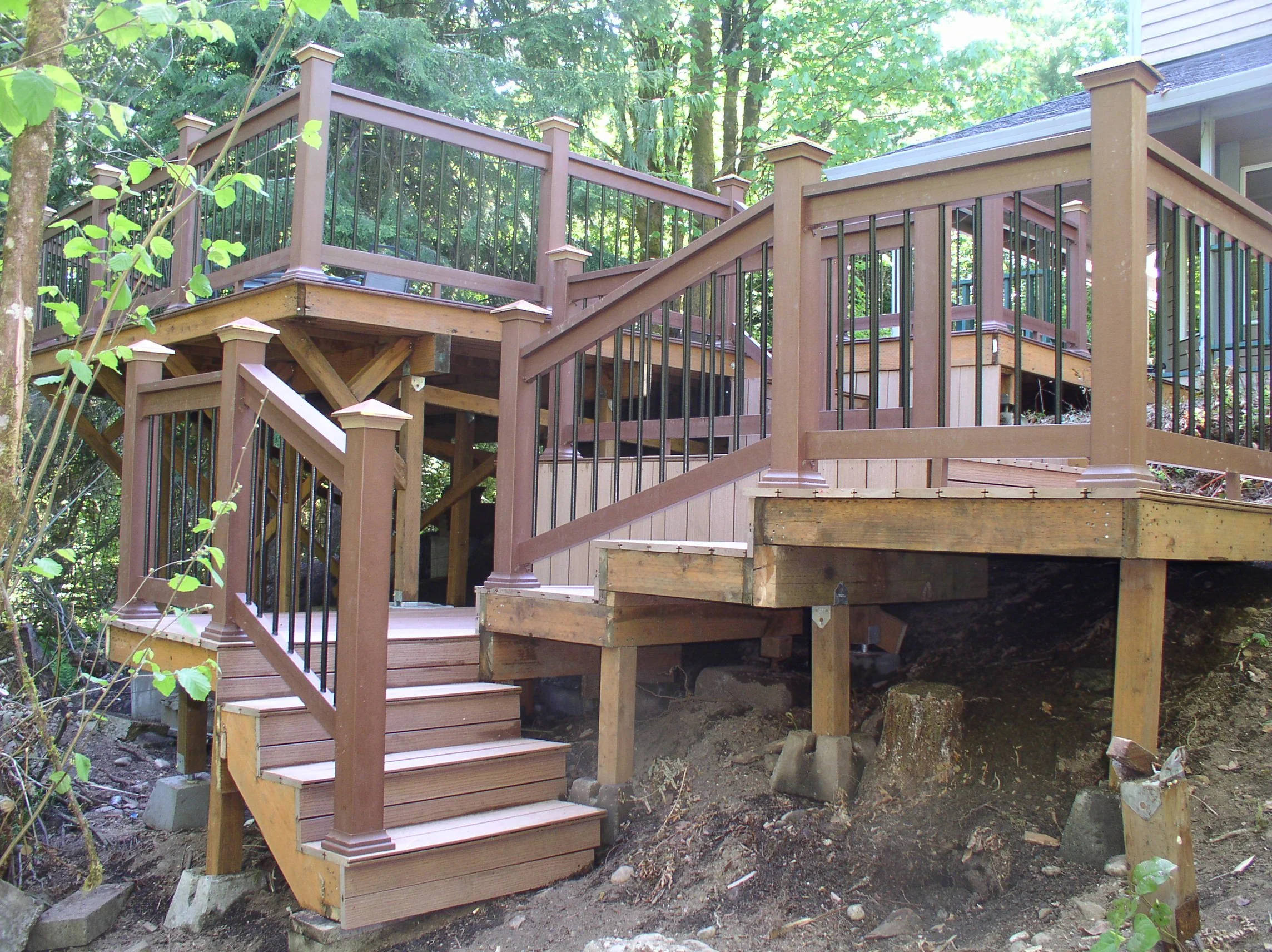 Newly built multi-level wooden deck with stairs and black metal railings, surrounded by trees and foliage.