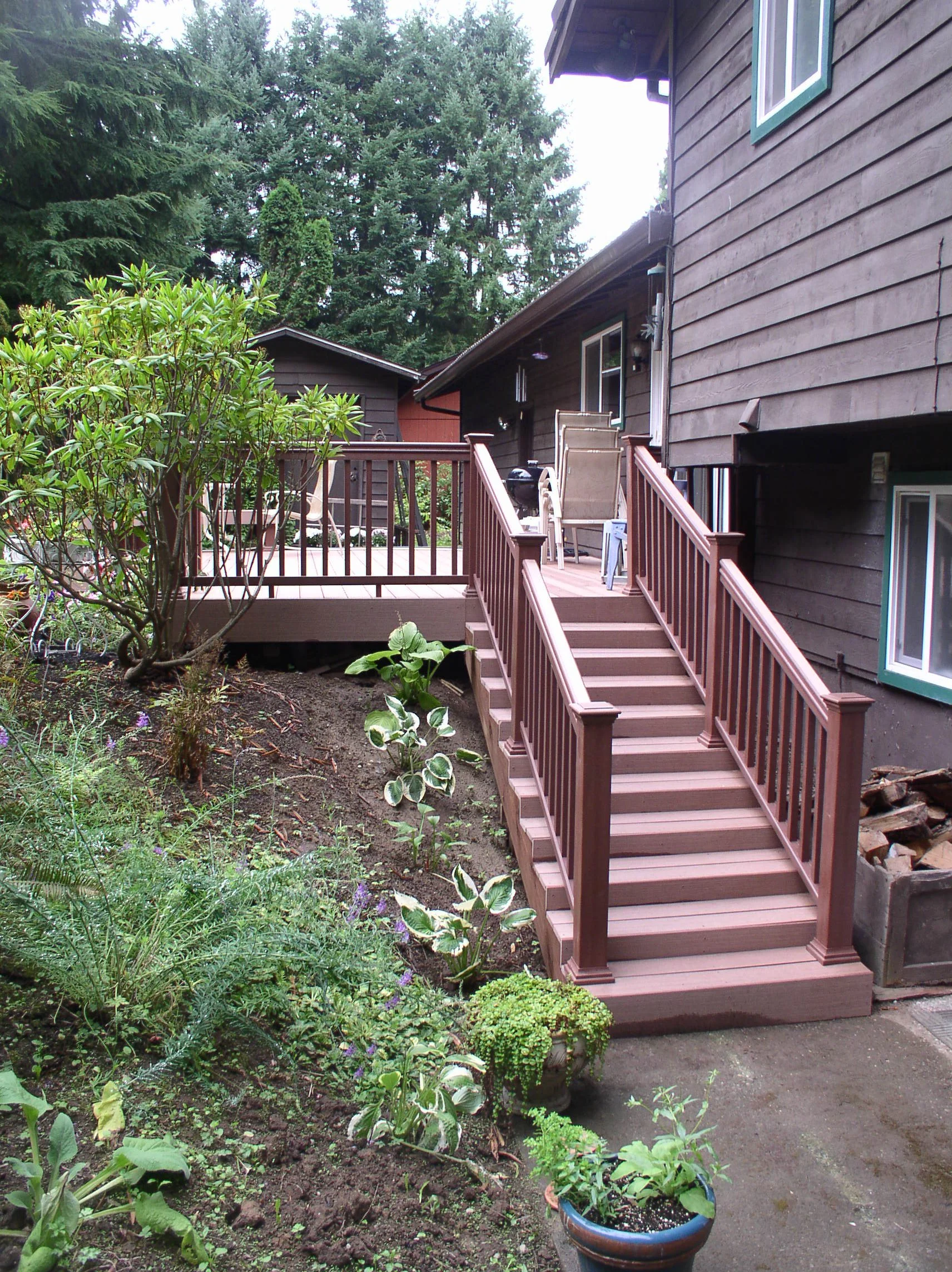 A backyard with a wooden deck accessible by a staircase, surrounded by greenery, plants, and trees.