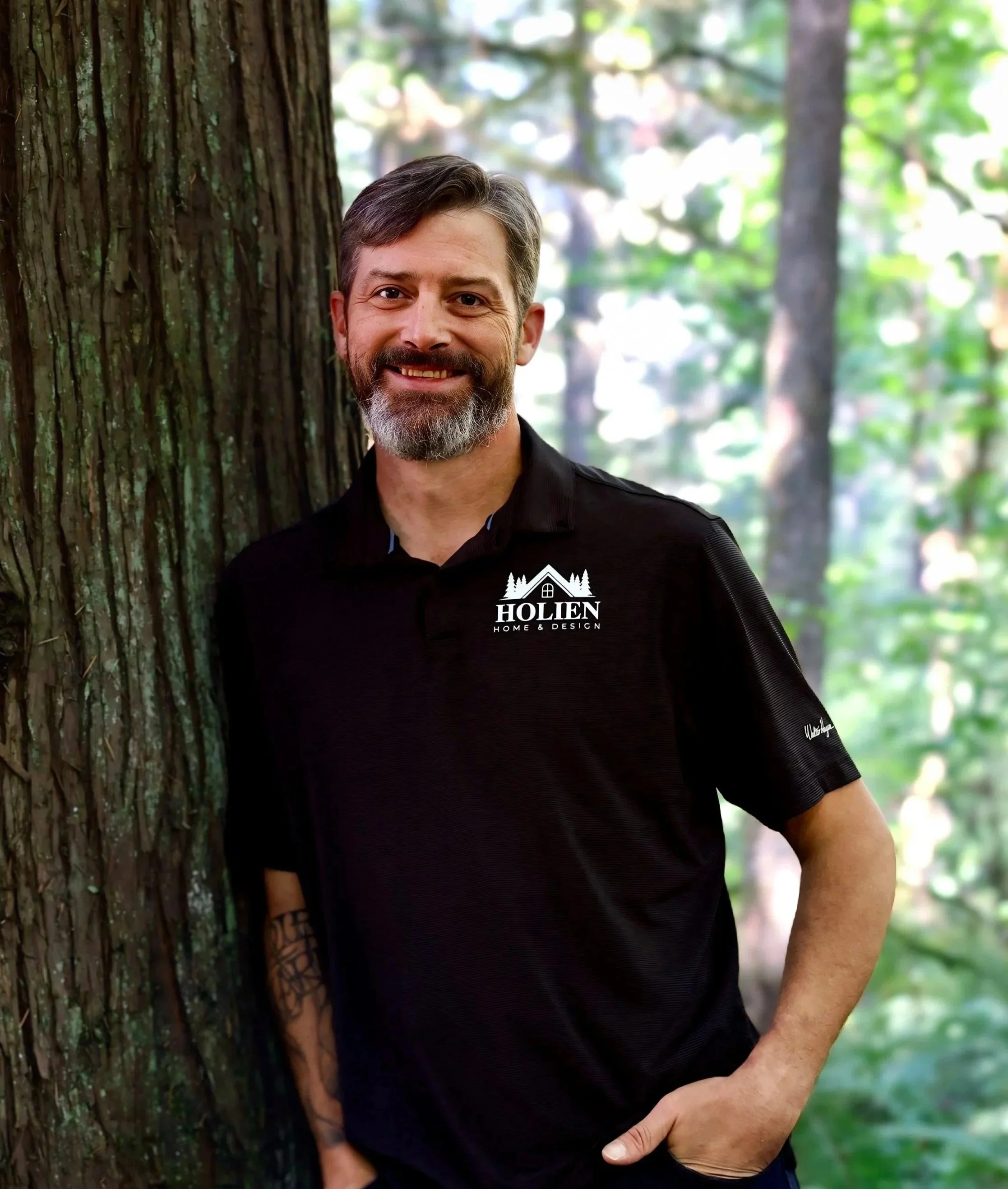 A man with a beard and mustache leaning against a tree in a forest, wearing a black polo shirt with the logo 'HOLIEN Home & Design' on it.
