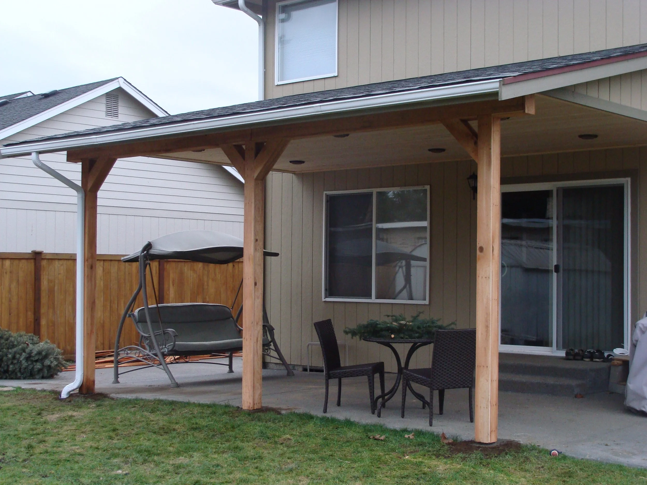 Backyard patio with a swing bench, a round table with two chairs, and a small Christmas tree, adjacent to a house with beige siding and sliding glass door.