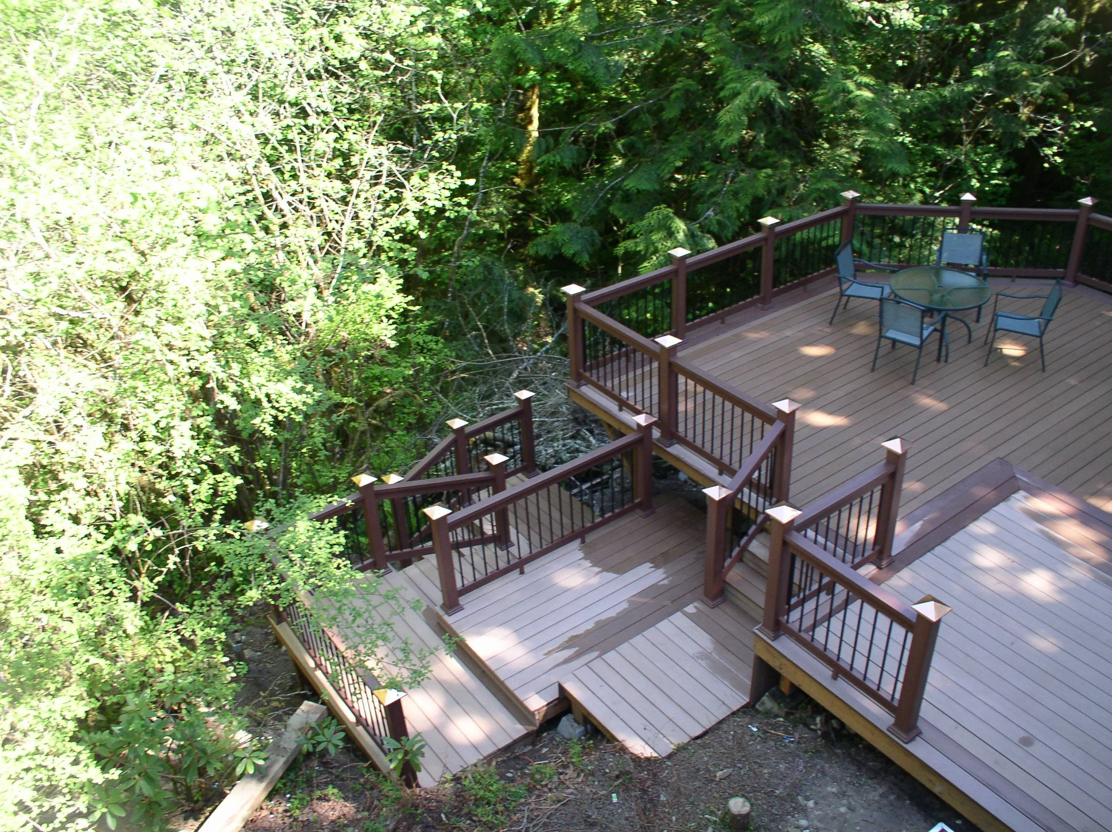 A wooden deck with outdoor furniture, including a round glass table and four chairs, situated among green trees and foliage.