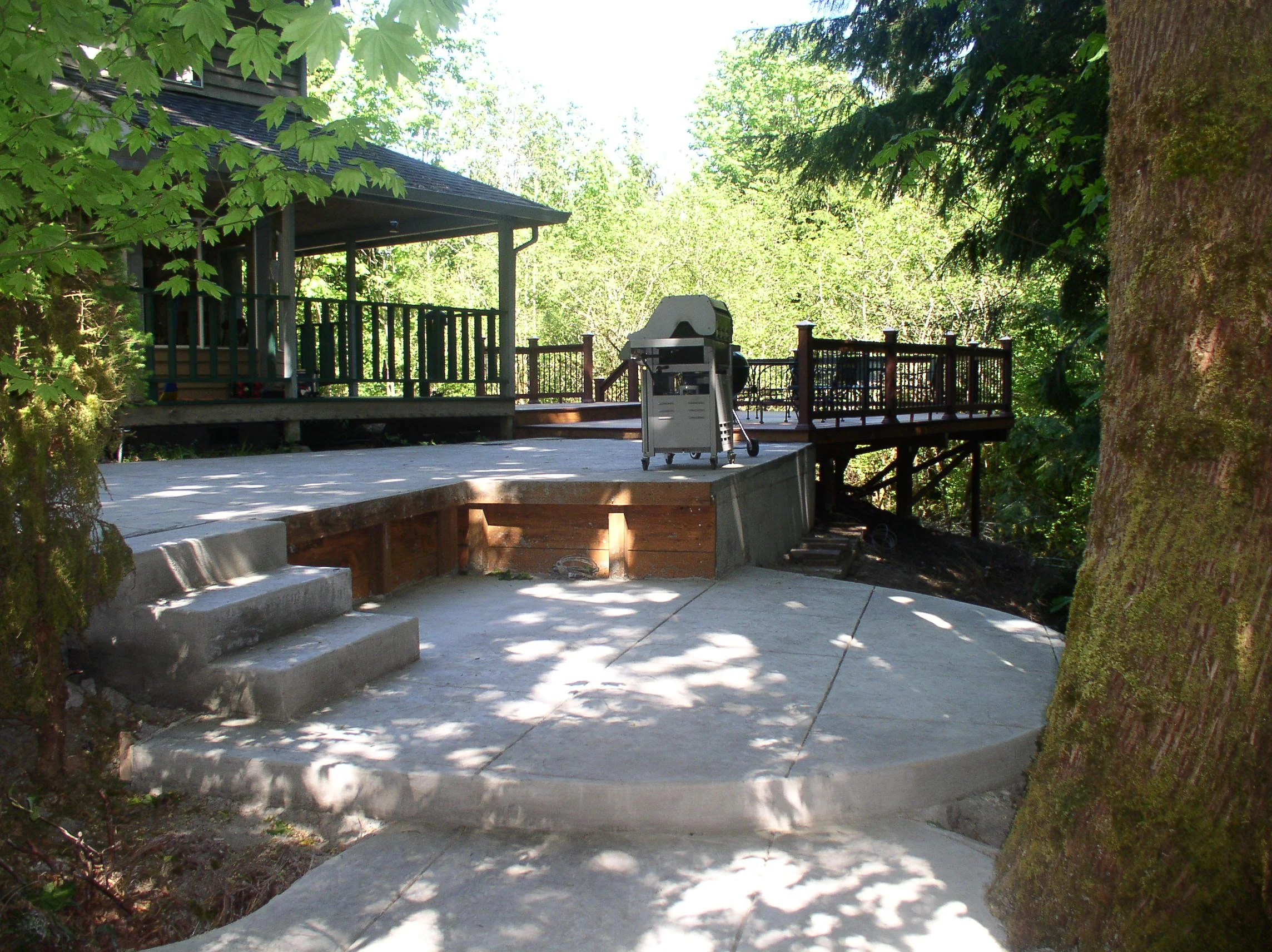 A newly constructed backyard patio with concrete step stairs, a wooden deck, a barbecue grill, and surrounded by green trees.