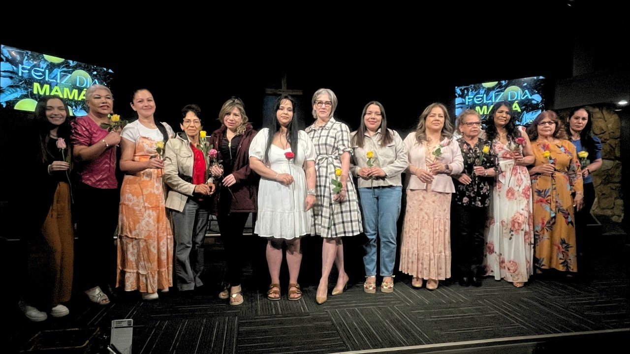 Group of women standing together on stage holding yellow and pink roses, celebrating Mother's Day with screen displays in the background