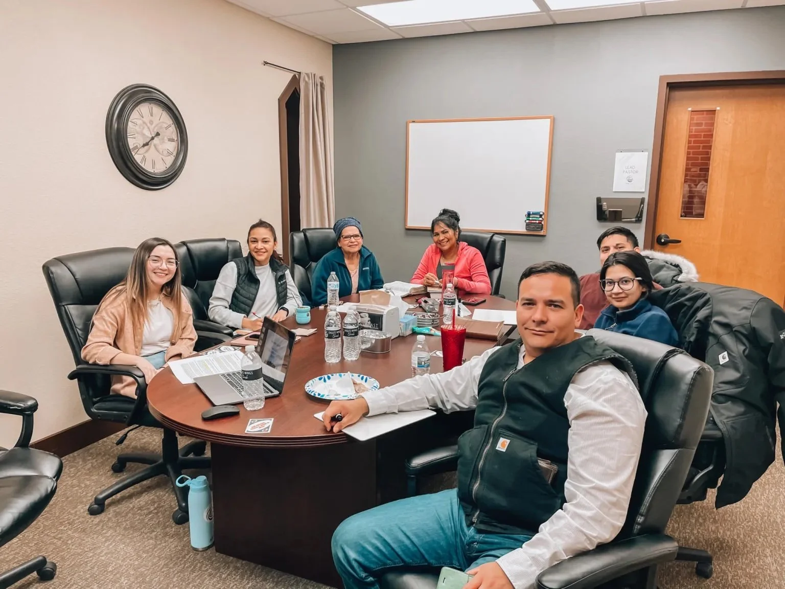 Seven diverse people sitting around a conference table in a meeting room, smiling at the camera, with laptops, water bottles, and papers on the table.