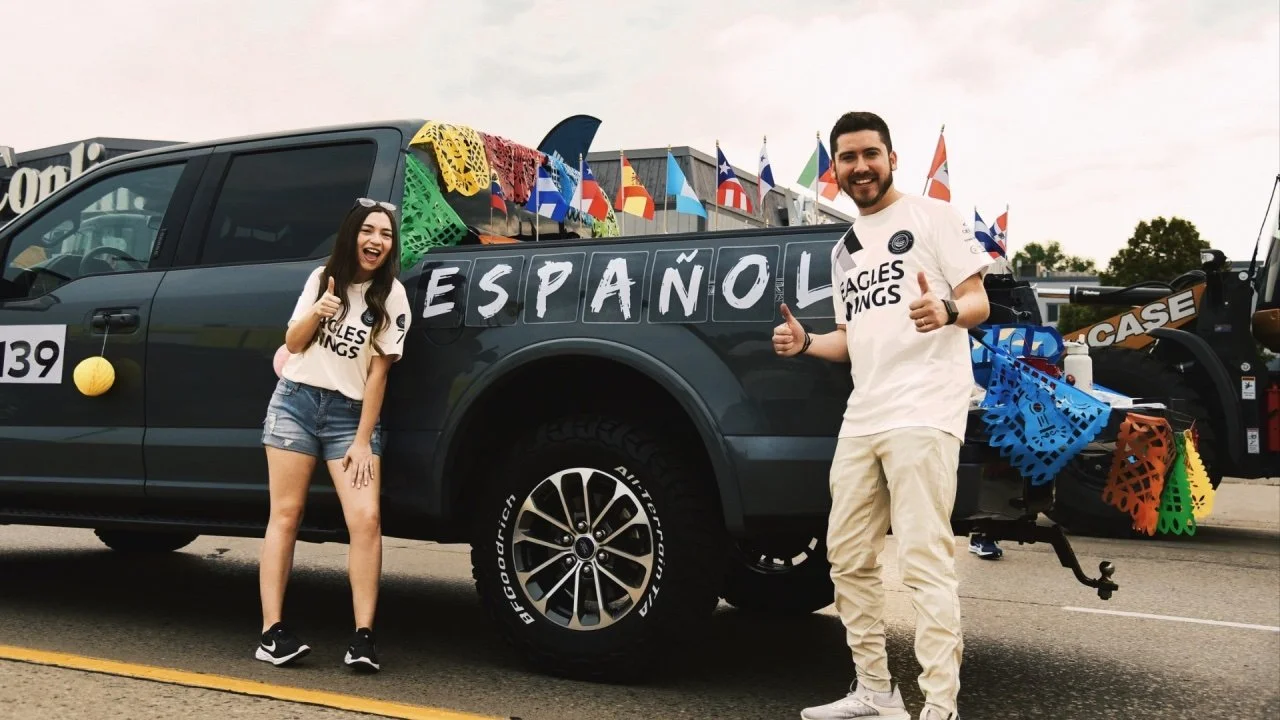 Two people smiling and giving thumbs up next to a decorated pickup truck with colorful flags and banners, with the word 'ESPAÑOL' on its side.