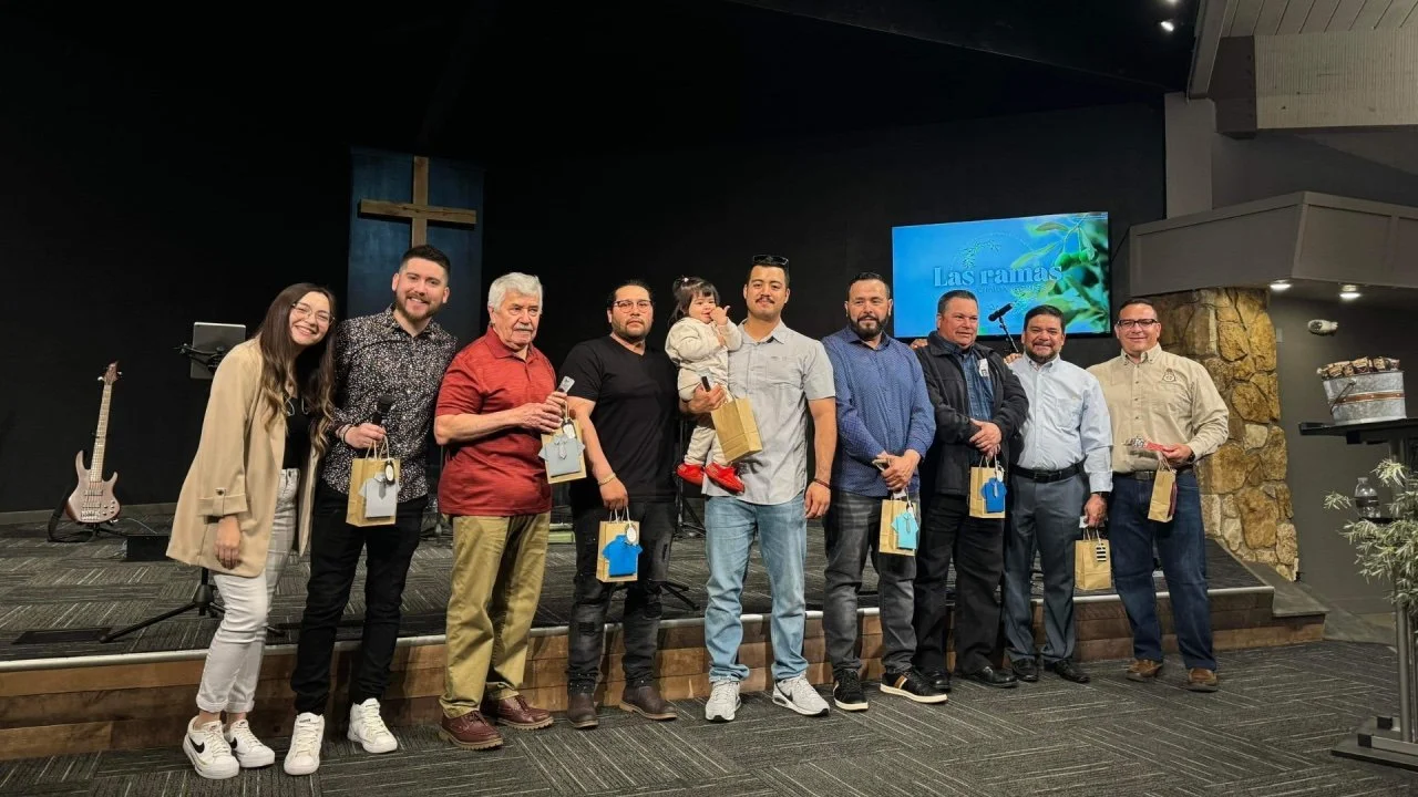 Group of ten people, including one holding a child, standing on a stage with a cross in the background, holding gift bags, in a church or religious venue.