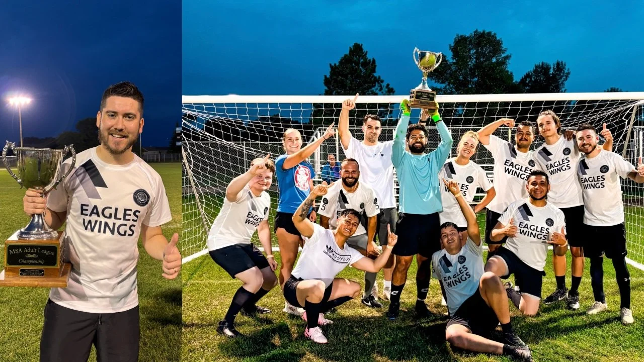 A soccer team celebrating a victory on the field at night, with some players wearing white jerseys that say "Eagles Wings" and holding a trophy, and one player holding a larger trophy.