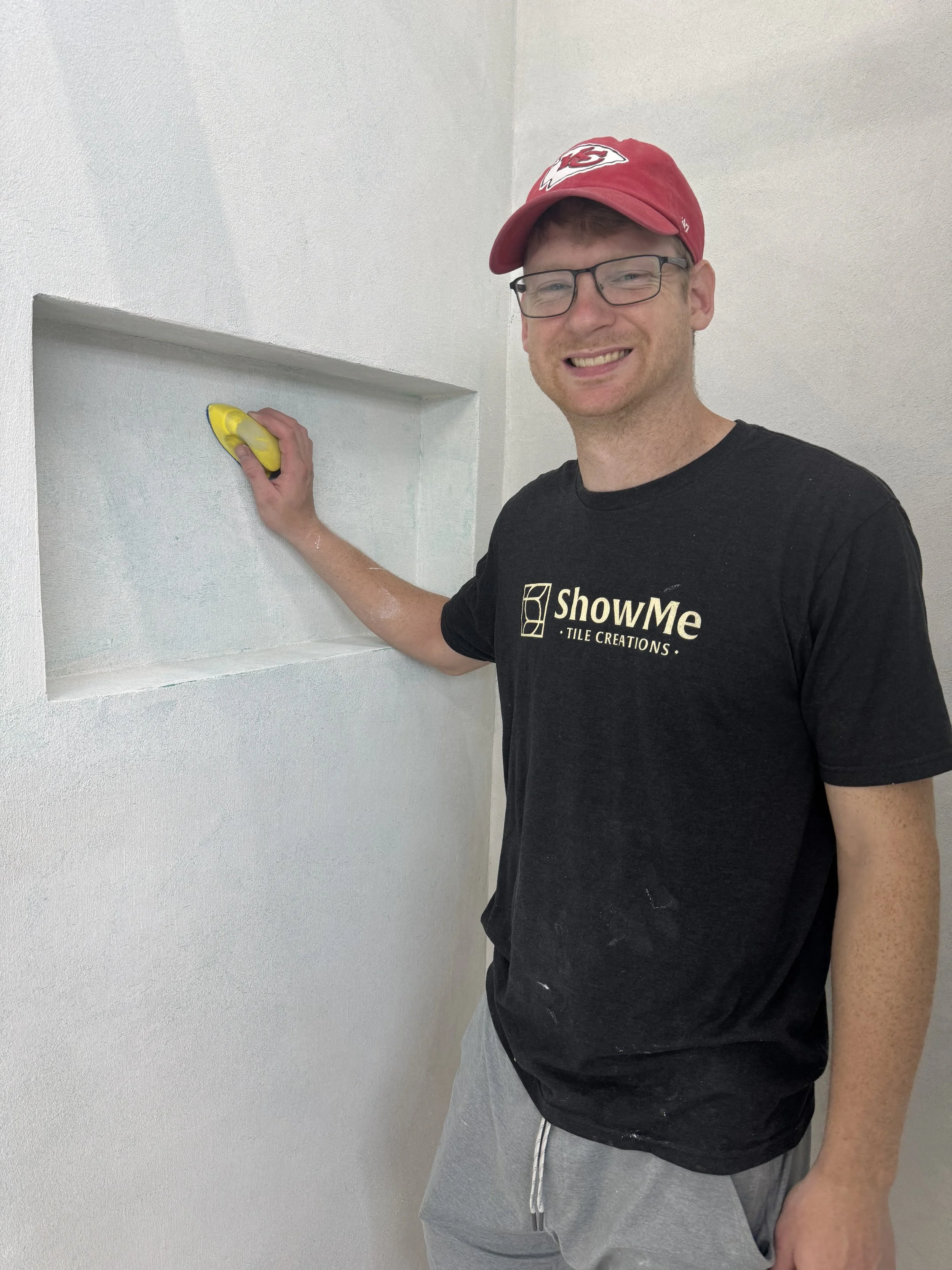 A man smiling at the camera while sanding a microcement recessed wall niche with a yellow sanding pad. He is wearing glasses, a red Kansas City Chiefs cap, a black T-shirt with the logo 'ShowMe Tile Creations,' and light gray shorts.
