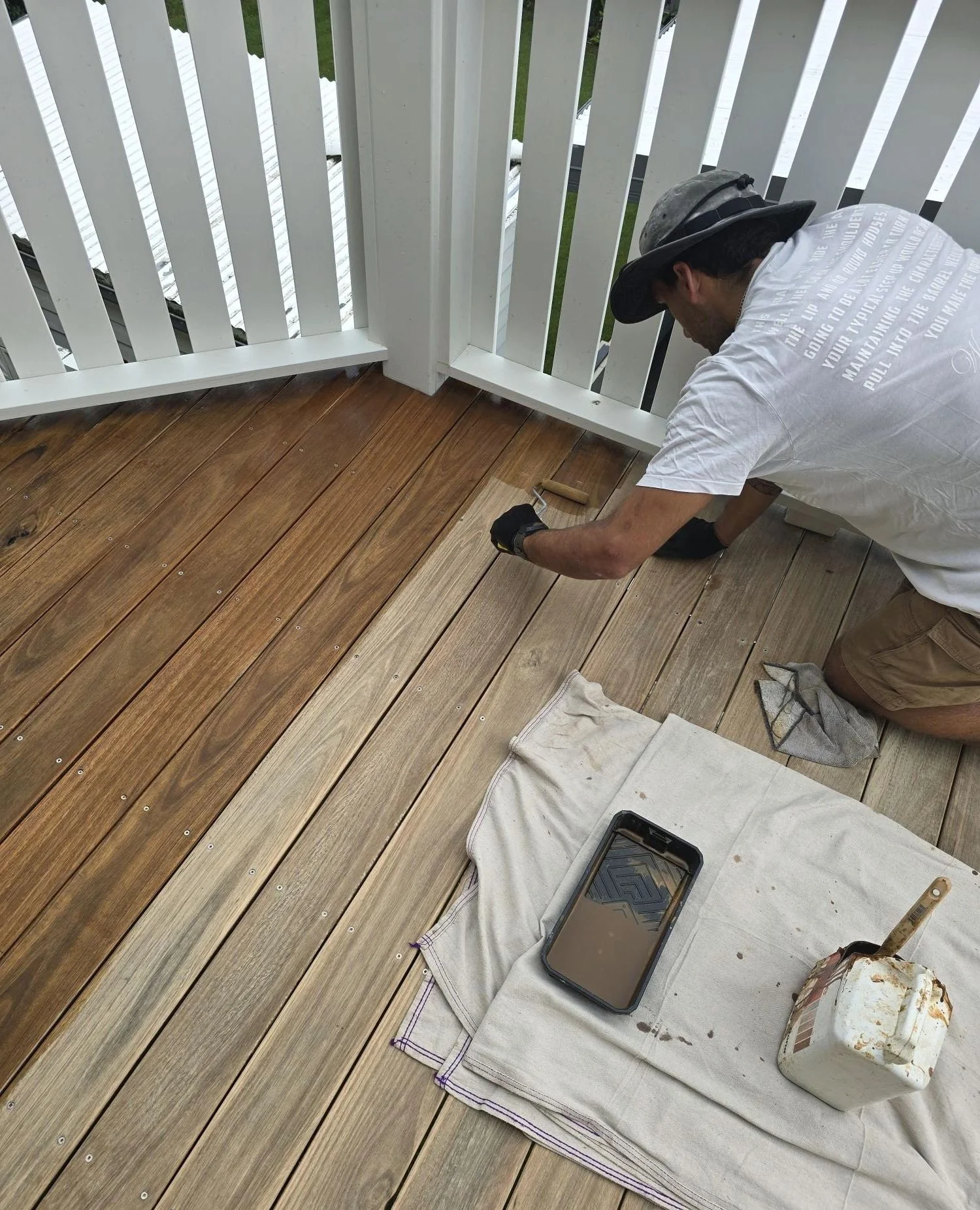A man kneeling on a wooden porch, working on installing or refinishing the wood flooring. He is using a small tool, with a paint roller nearby, and has a smartphone and a container on a cloth on the porch.