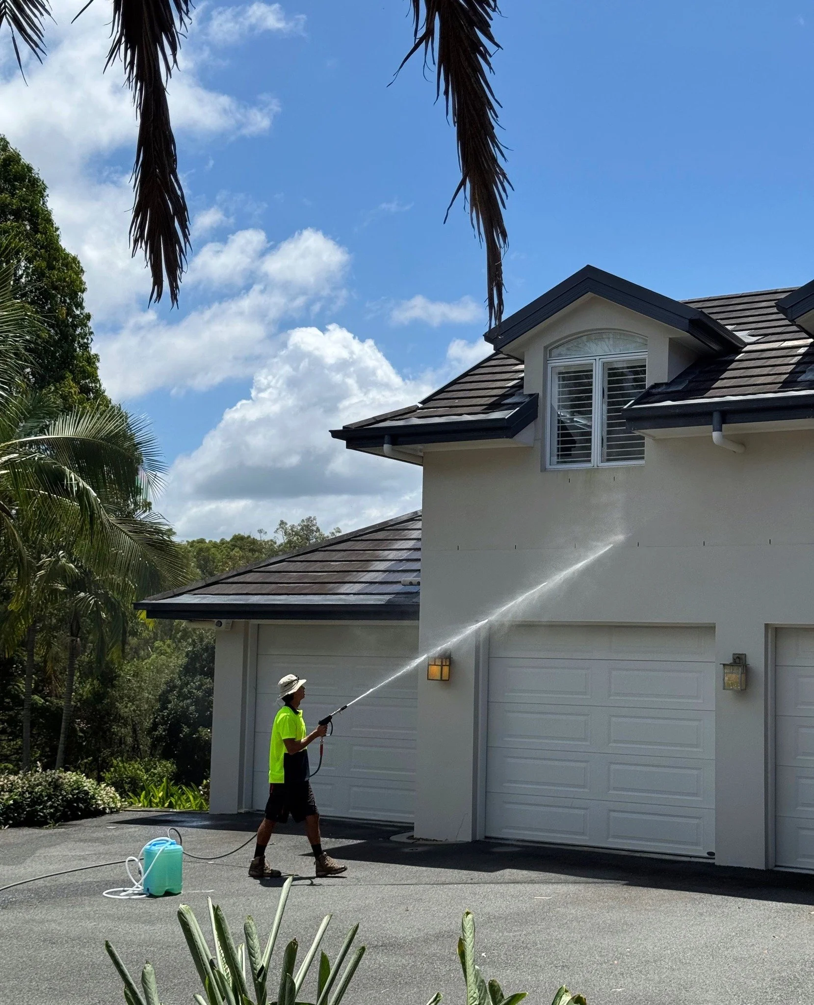 A person wearing a neon yellow shirt, dark shorts, and a wide-brimmed hat is spraying water on a house's exterior wall using a hose, with the house's garage doors visible and trees in the background under a partly cloudy sky.