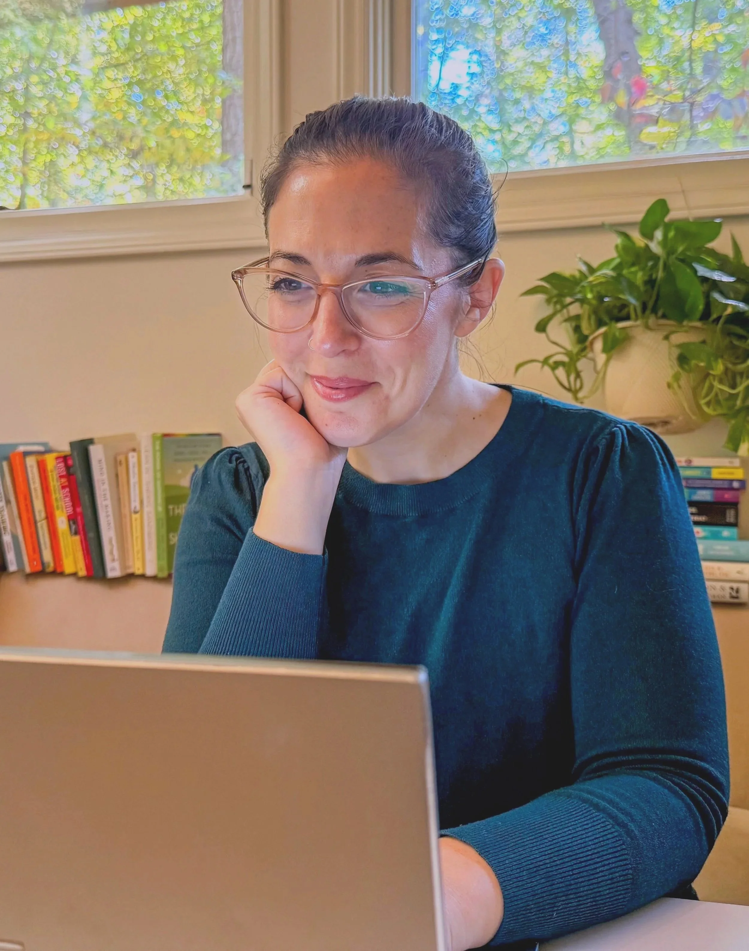A woman with glasses working on a laptop in a room with a bookshelf and a potted plant, with a window showing trees outside.