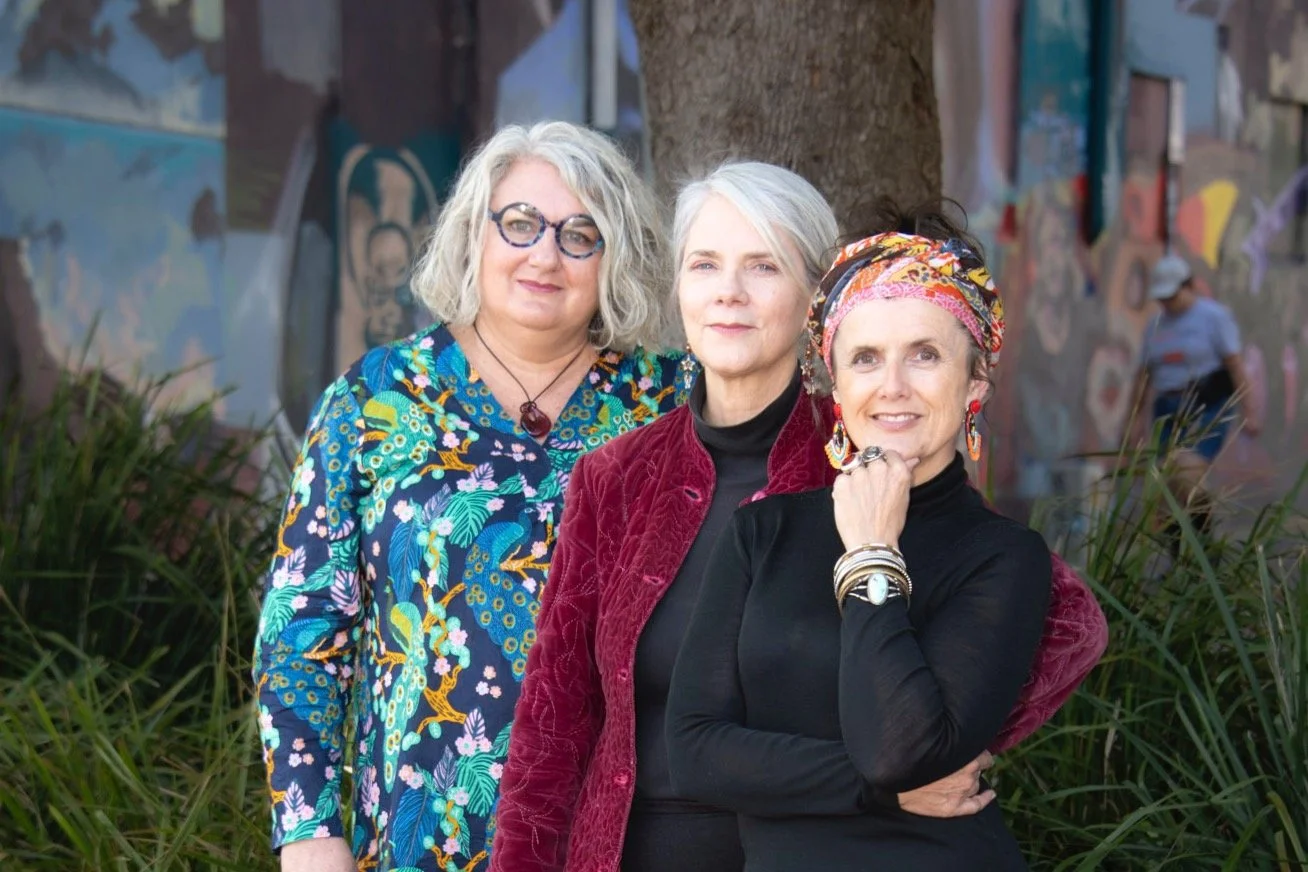 Three women standing outdoors in front of a mural and foliage, posing for a photo. One woman wears a colorful patterned blouse and glasses, another wears a dark turtleneck and red jacket, and the third woman wears a black top with a colorful headscarf and jewelry.