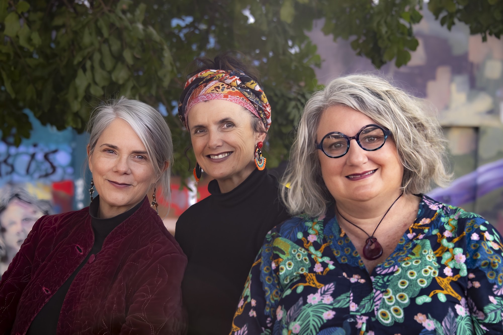 Three women musicians in a band smiling and posing outdoors, with trees and a cityscape in the background.