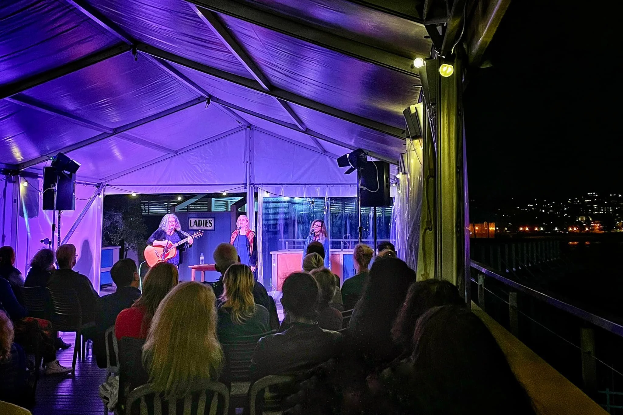 A group of women vocalists and musicians performing on a stage under purple lighting inside a tent at night, with audience members seated watching.