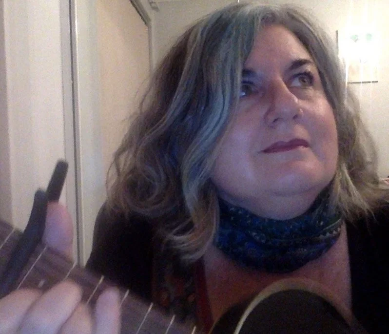 A woman with wavy, shoulder-length hair playing an acoustic guitar, wearing a dark top and a patterned neck scarf, in a room with a window and a white door. Christina Mimmocchi choir director, musician, singer songwriter