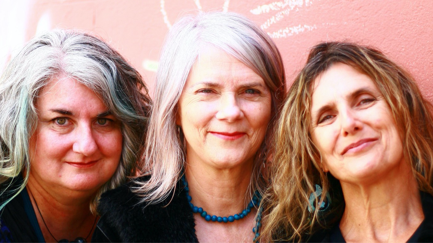 Three women standing close together outdoors, smiling, with a pink wall in the background