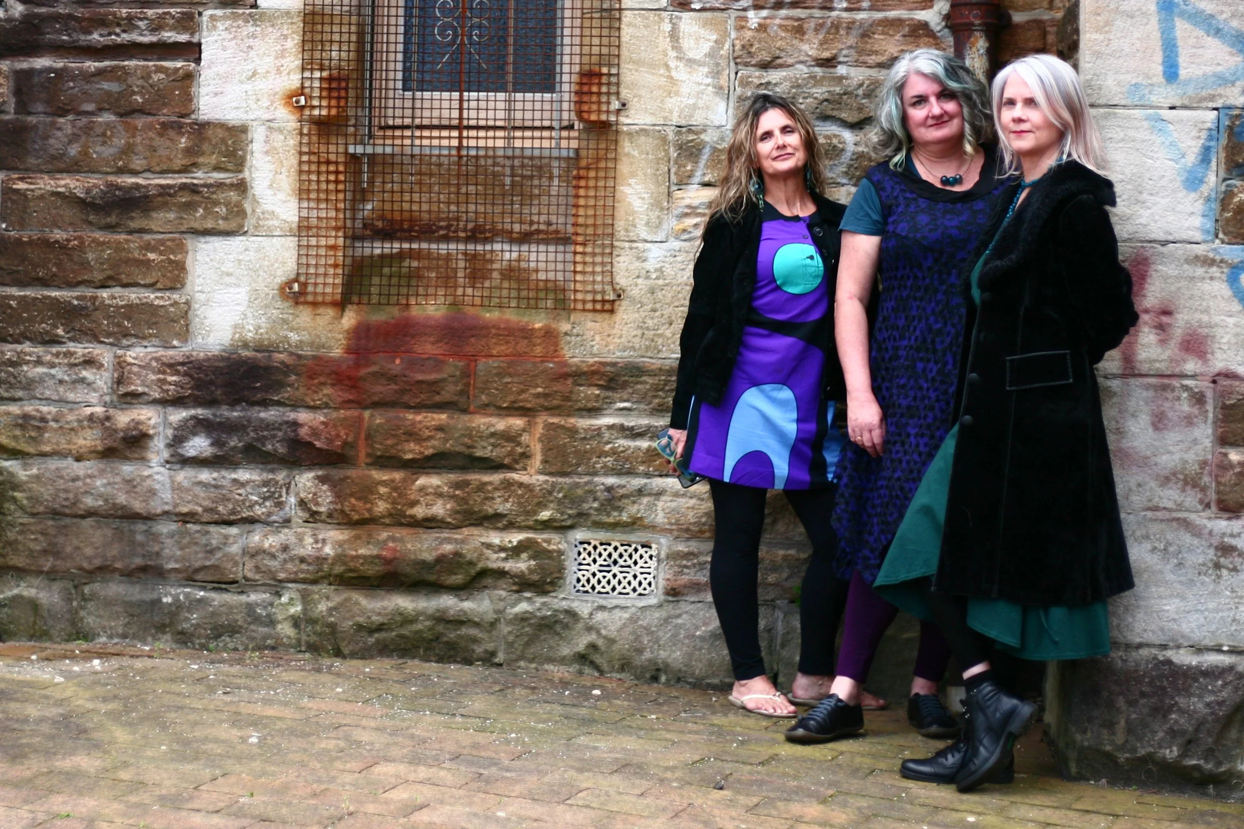 Three women standing in front of a graffiti-covered brick wall, wearing stylish clothing.